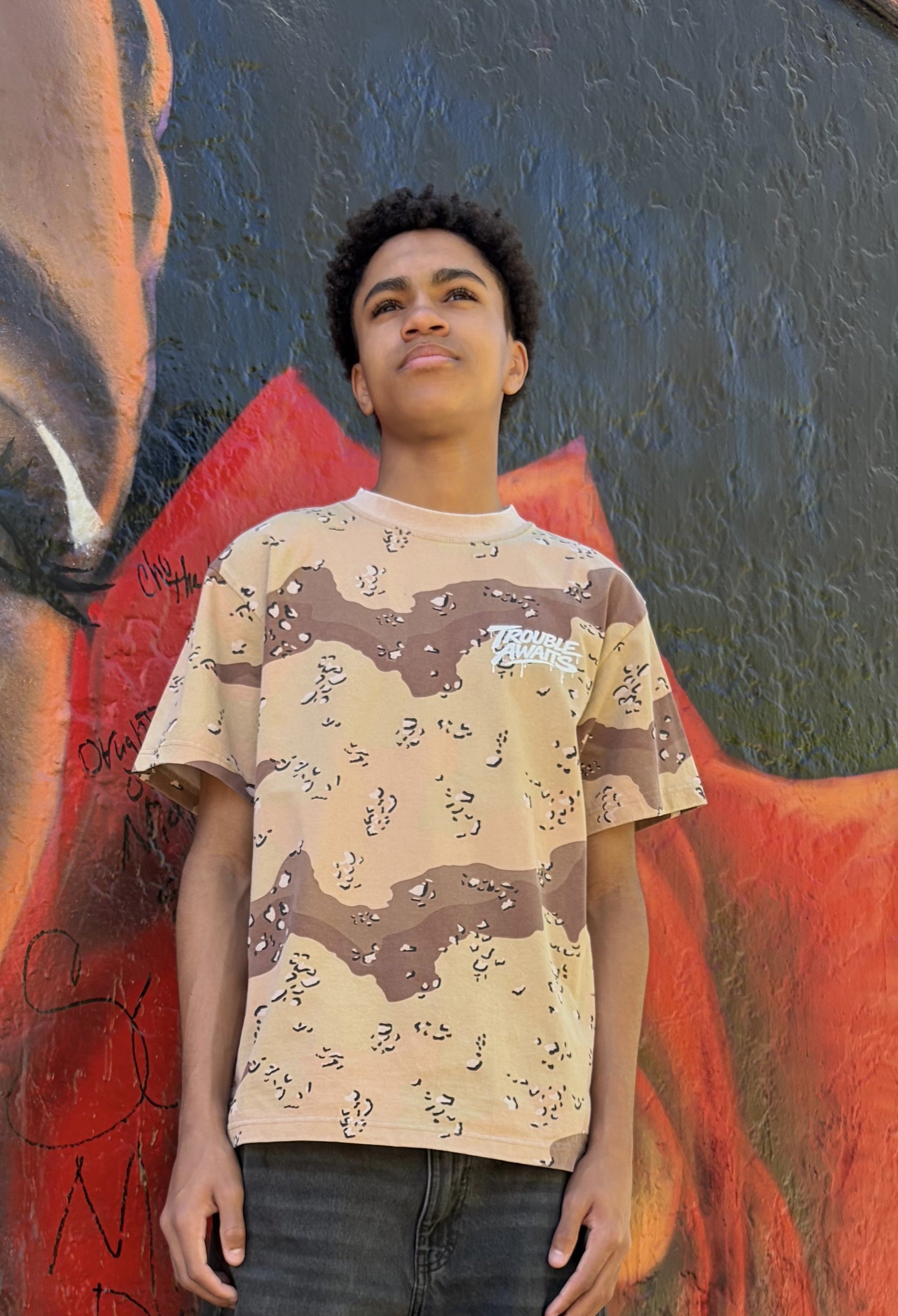A person stands outdoors in front of a textured wall covered in bold, colorful graffiti artwork. The background features dark gray and black tones with bright red and orange shapes, along with scattered tags and painted details.

The person is wearing an oversized, boxy short-sleeve T-shirt with a desert camouflage pattern in shades of tan, beige, and brown, with small darker speckles throughout. On the upper left chest, there is a small white “Trouble Awaits” logo in a graffiti-style font 