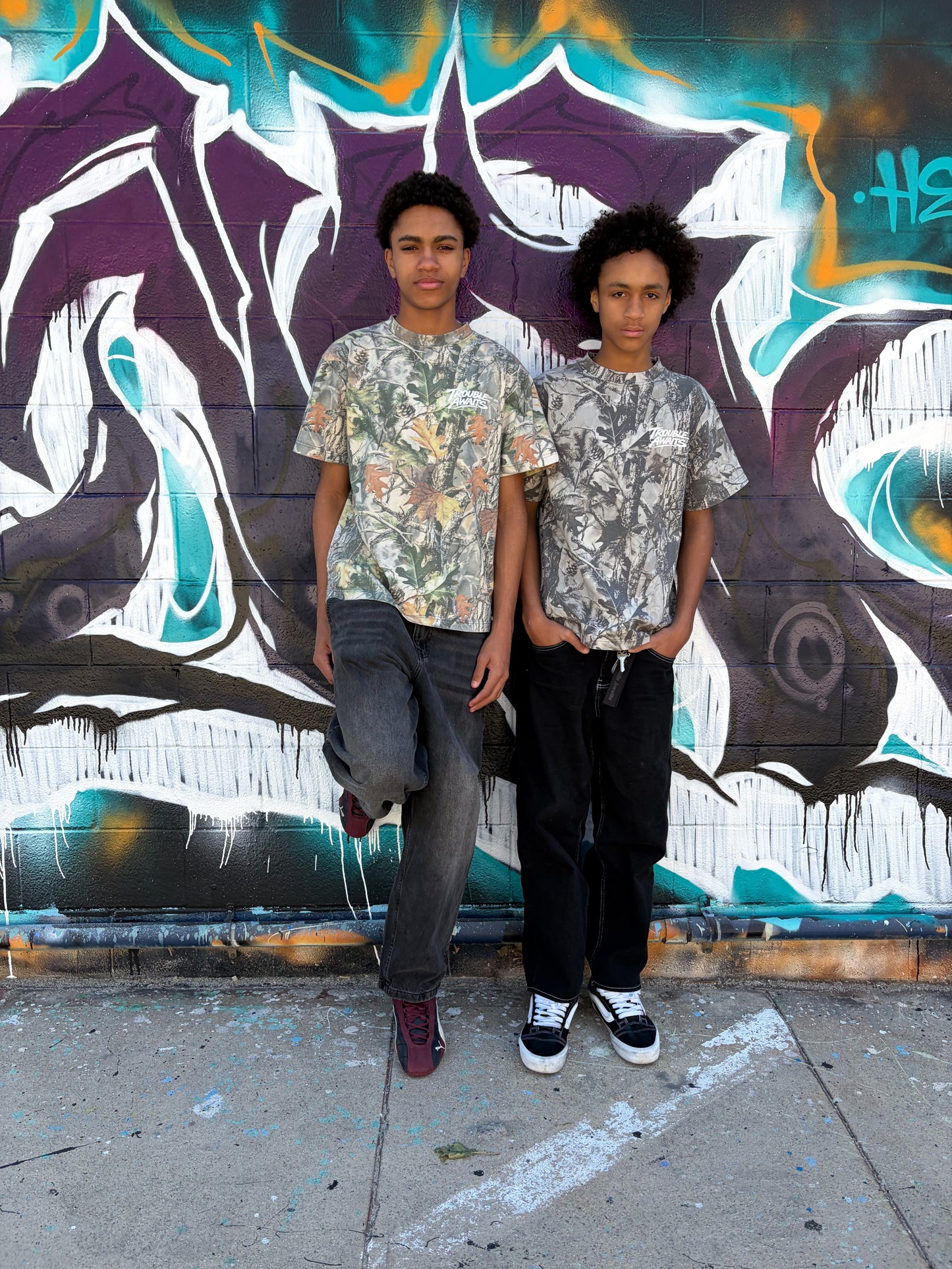 Two people stand side by side in front of a brick wall covered in graffiti art. The mural behind them features bold shapes and layered colors, including purple, teal, white, and hints of orange, creating a textured urban backdrop.

Both individuals face the camera with neutral expressions and relaxed postures. They appear to be of similar age and height, with short, curly hair.

They are each wearing oversized, boxy short-sleeve T-shirts featuring a realistic woodland camouflage pattern. 