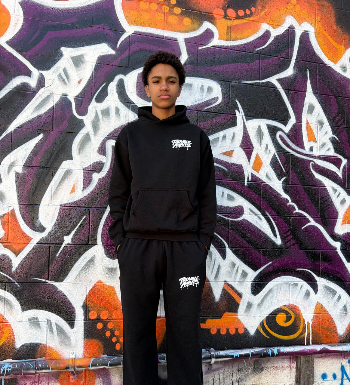 Person standing facing the camera wearing a black hoodie and matching black sweatpants set. The hoodie features a small white “Trouble Awaits” logo on the left chest, and the sweatpants have the same logo on the upper thigh. The person has short curly hair and a neutral expression, with hands placed in the front pockets of the pants. They are standing in front of a vibrant graffiti mural painted on a brick wall, featuring abstract shapes in white, purple, black, and orange.