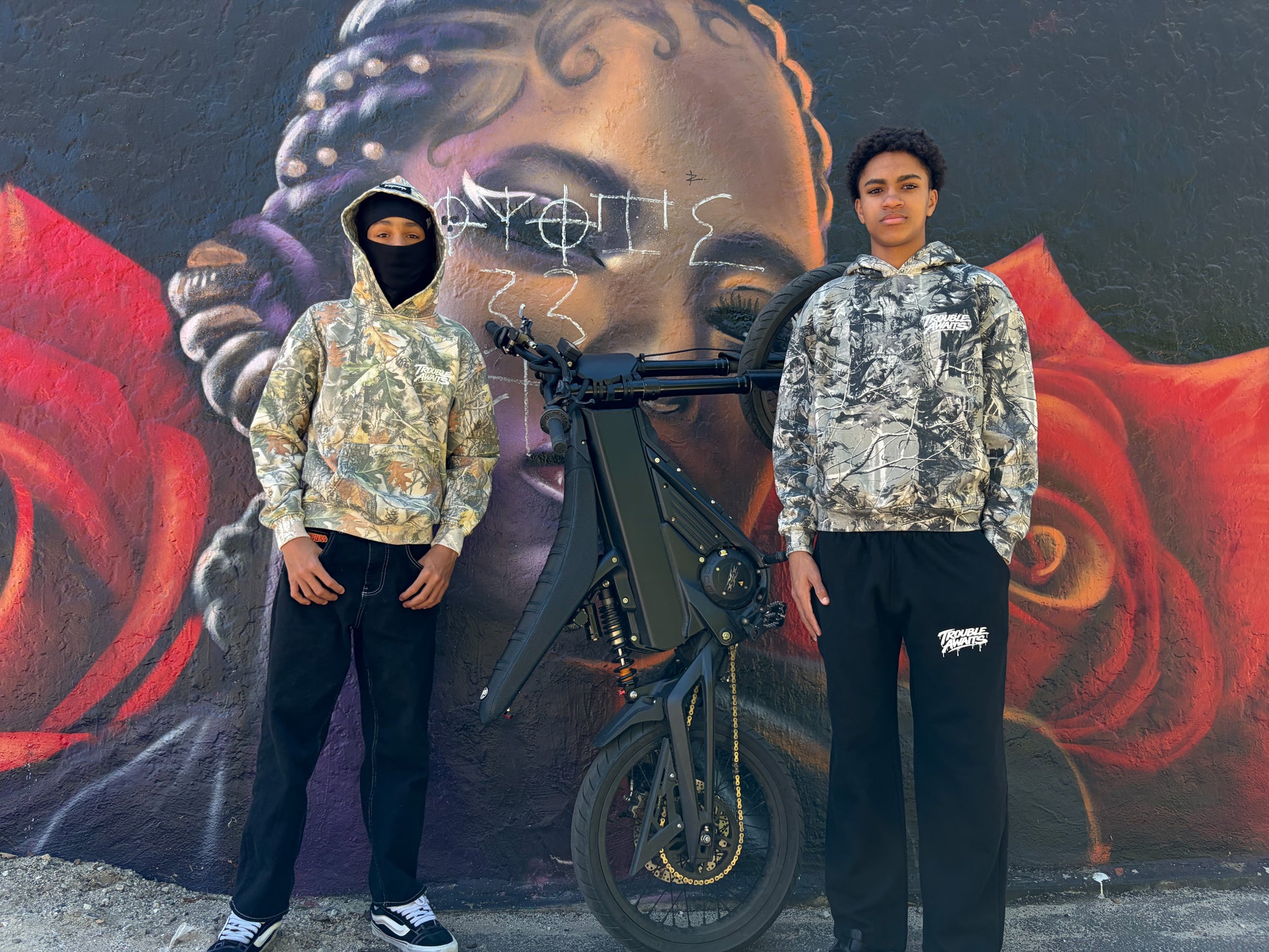 This image shows two people standing outdoors in front of a large mural painted on a textured wall. The mural features a stylized portrait of a face with braided hair on the left side, along with bold red and dark background elements. Positioned between the two individuals is a black electric-style bike with a visible frame, handlebars, and front wheel angled slightly upward.
They are both wearing camo hoodies with Trouble Awaits white logo on left chest.