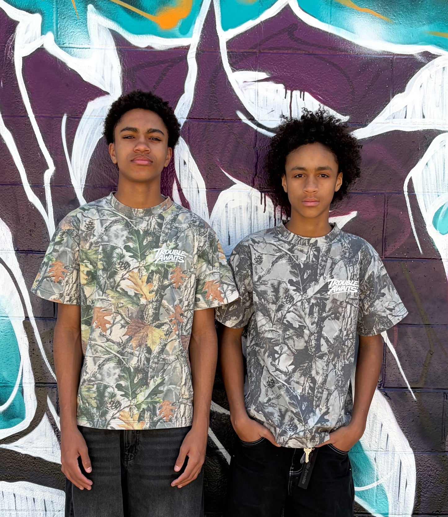 Two people stand side by side in front of a brick wall covered in graffiti art. The mural behind them features bold shapes and layered colors, including purple, teal, white, and hints of orange, creating a textured urban backdrop.

Both individuals face the camera with neutral expressions and relaxed postures. They appear to be of similar age and height, with short, curly hair.

They are each wearing oversized, boxy short-sleeve T-shirts featuring a realistic woodland camouflage pattern. 
