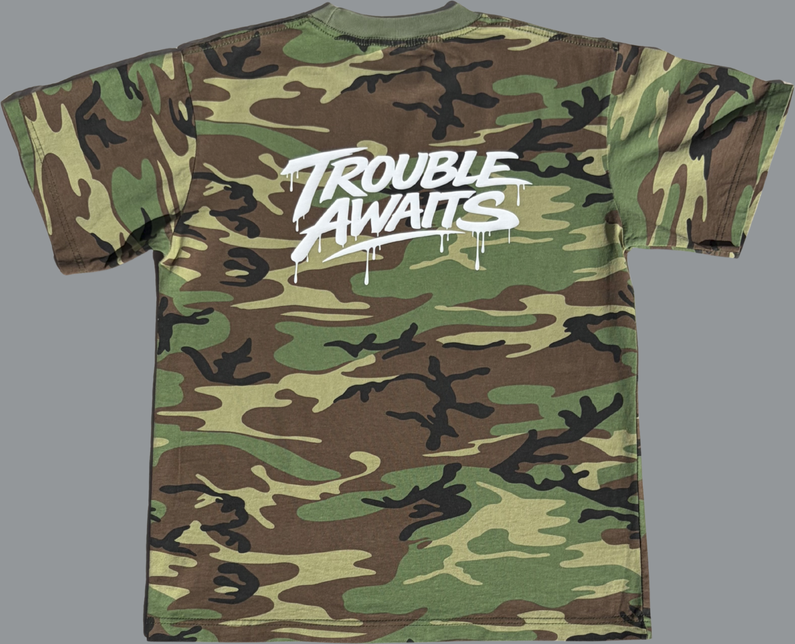 This is the back view of a short-sleeve T-shirt featuring a classic woodland camouflage pattern in shades of green, brown, tan, and black. Centered across the upper back is a bold white graphic that reads “Trouble Awaits” in a stylized graffiti-inspired font. The lettering has a slightly dripping paint effect, giving it a streetwear, hand-painted look. The shirt has a standard crew neck and relaxed fit, with the camo pattern covering the entire garment. The high-contrast white text stands out sharply 