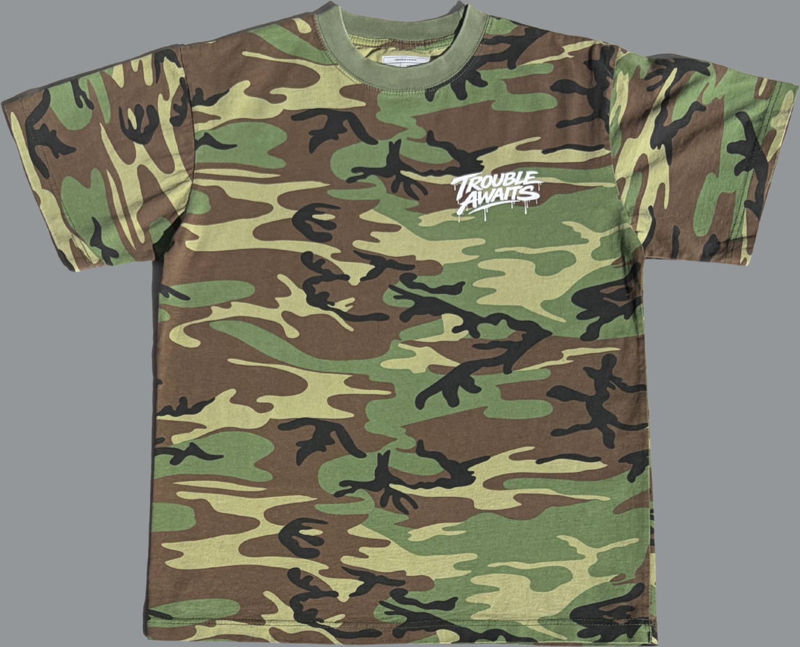 This is the front view of a short-sleeve T-shirt featuring a classic woodland camouflage pattern in shades of green, brown, tan, and black. The shirt has a crew neckline with a matching green ribbed collar. On the upper left chest, there is a small white graphic that reads “Trouble Awaits” in a graffiti-style font with a subtle dripping paint effect. The rest of the shirt is covered entirely in the camo print, giving it a rugged, military-inspired look. The fit appears relaxed and casual.