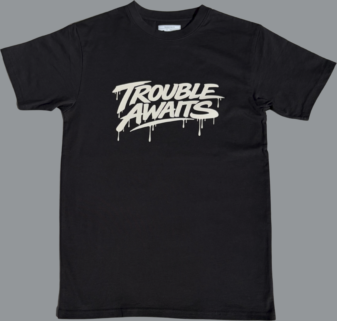 Black short sleeve t-shirt featuring a large tan graphic centered on the chest. The design reads “Trouble Awaits” in bold stylized lettering with a dripping paint effect extending from several of the letters. The rest of the shirt is plain black with no additional visible graphics.