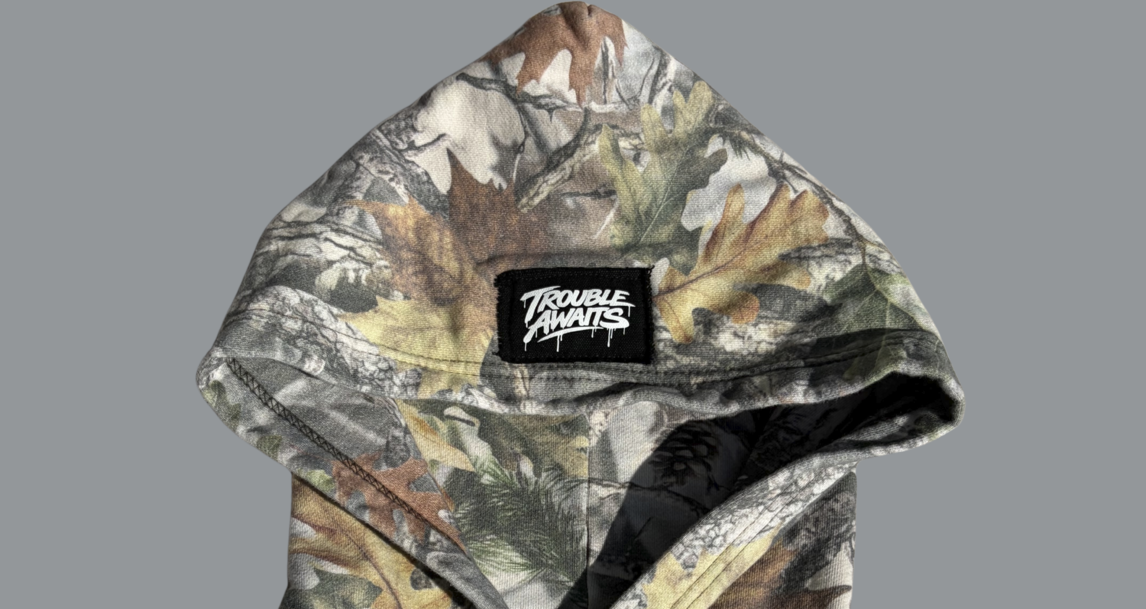 Close-up view of the hood on a camouflage zip-up hoodie in a woodland pattern of muted greens, browns, and beige. Centered at the back of the hood is a black rectangular woven patch with white “Trouble Awaits” branding in a graffiti-style font. The fabric shows detailed leaves and branches, and the hood opening is slightly parted, revealing a darker interior lining.