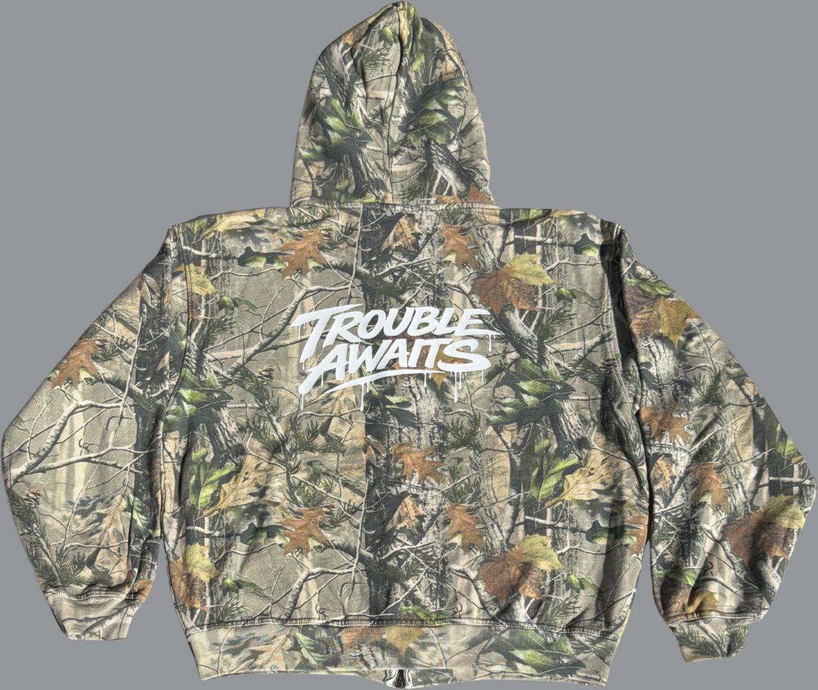 Back view of an oversized, boxy zip-up hoodie with dropped shoulders in a woodland camouflage pattern. The design features layered branches, leaves, and tree bark in muted greens, browns, and beige tones. Centered across the upper back is a large white “Trouble Awaits” graphic in a bold, graffiti-style font with subtle drip detailing. The hoodie has a roomy hood, relaxed wide sleeves, and ribbed cuffs and hem, emphasizing its loose, streetwear-inspired fit.