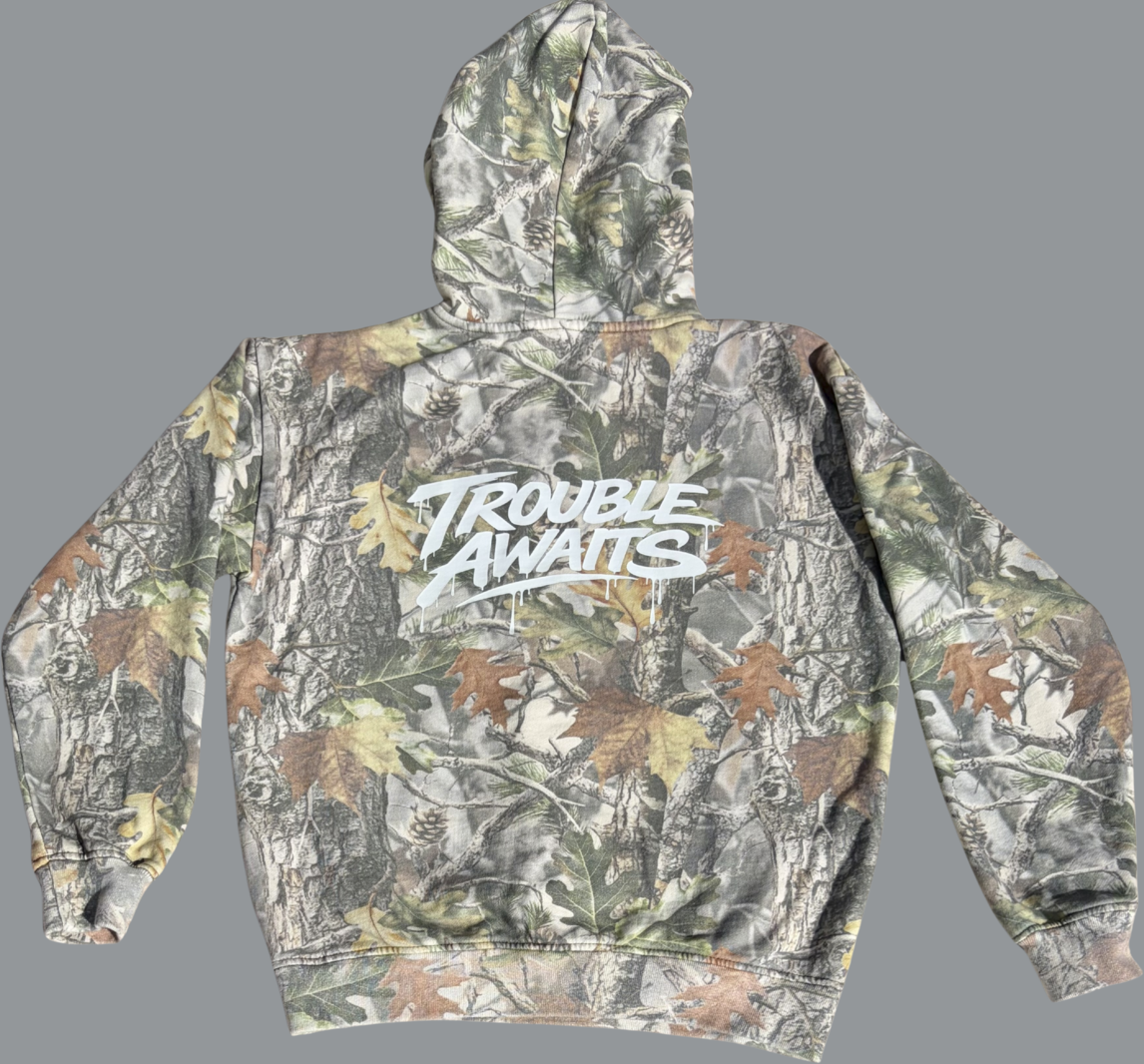 This image shows the back view of a pullover hoodie laid flat against a neutral gray background. The hoodie features a realistic forest camouflage pattern made up of tree bark textures, branches, and scattered leaves in shades of green, brown, tan, and gray. Across the upper back is a large white “Trouble Awaits” logo printed in a bold, graffiti-inspired script with paint-drip effects extending downward from the lettering. The hood is visible at the top, and the garment has long sleeves with ribbed cuffs