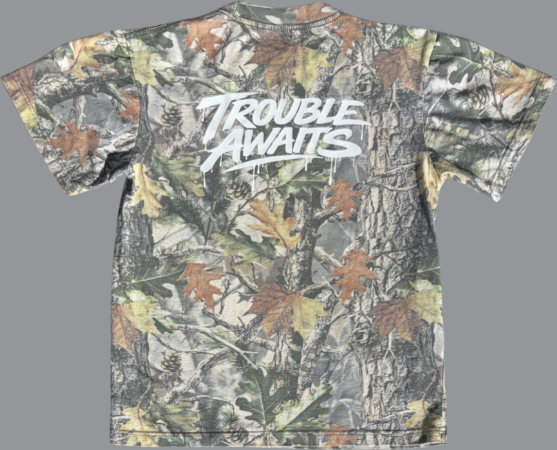 A back view of an oversized, boxy short-sleeve T-shirt displayed flat against a neutral gray background. The shirt features a realistic woodland camouflage pattern made up of tree bark textures, branches, and scattered leaves in shades of green, brown, tan, and muted gray.

Centered across the upper back is a large white “Trouble Awaits” graphic in a bold, graffiti-style script. The lettering includes dripping paint details beneath several strokes, giving it a street-art-inspired look 