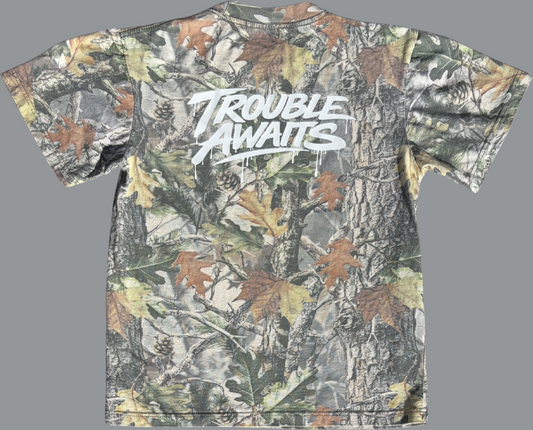 A back view of an oversized, boxy short-sleeve T-shirt displayed flat against a neutral gray background. The shirt features a realistic woodland camouflage pattern made up of tree bark textures, branches, and scattered leaves in shades of green, brown, tan, and muted gray.

Centered across the upper back is a large white “Trouble Awaits” graphic in a bold, graffiti-style script. The lettering includes dripping paint details beneath several strokes, giving it a street-art-inspired look 