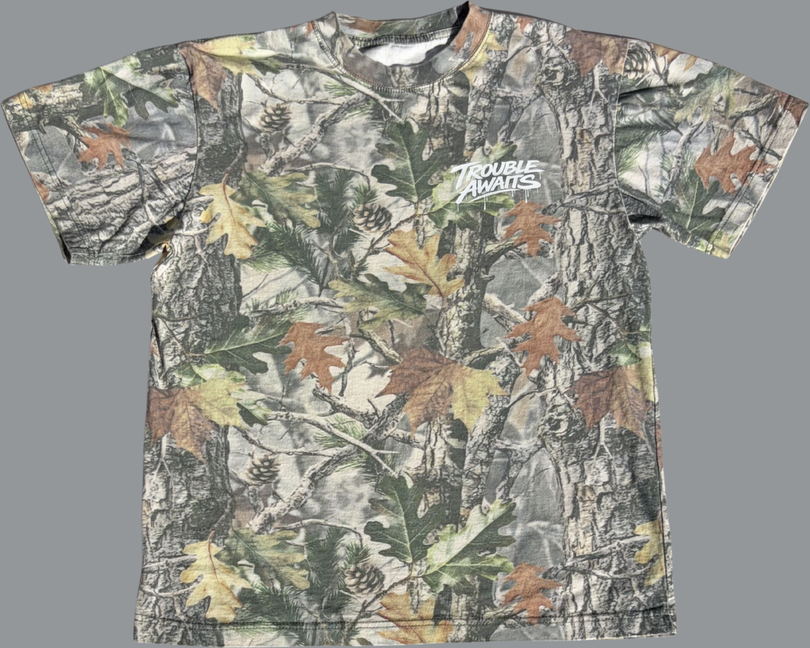 A front view of an oversized, boxy short-sleeve T-shirt displayed flat against a neutral gray background. The shirt features a realistic woodland camouflage pattern composed of tree bark textures, branches, and scattered leaves in shades of green, brown, tan, and muted gray.

On the upper left chest, there is a small white “Trouble Awaits” graphic in a graffiti-style script with subtle dripping paint details. The bright white logo stands out against the natural-toned camouflage pattern.


