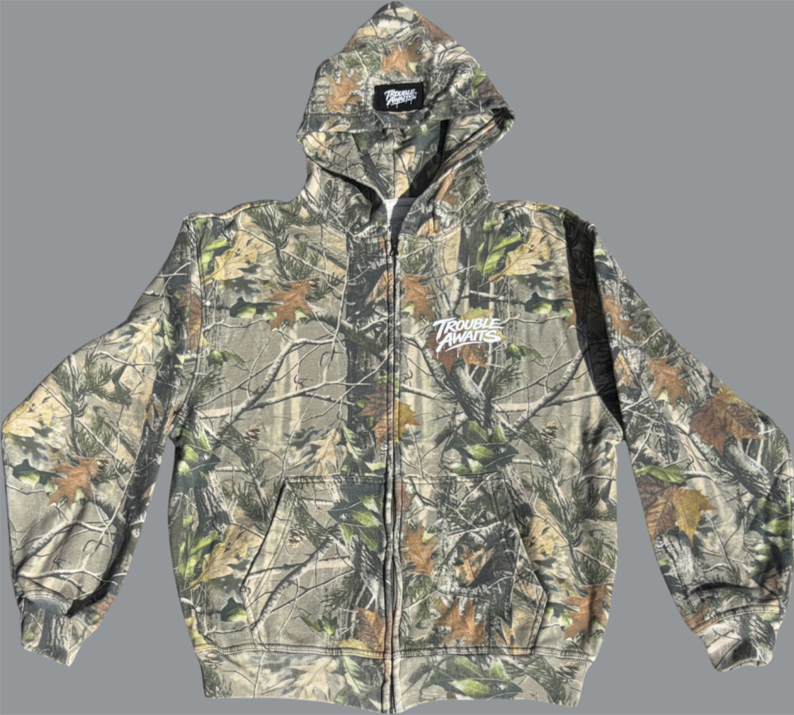 Front-facing image of an oversized, boxy zip-up hoodie with dropped shoulders in a woodland camouflage pattern. The print features layered branches, leaves, and tree bark in muted greens, browns, and beige tones. The hoodie has a full-length front zipper, a large hood, ribbed cuffs and hem, and a split kangaroo pocket at the front. A white “Trouble Awaits” logo is printed on the left chest. Inside the hood, a black woven label with white branding is visible at the back of the collar.