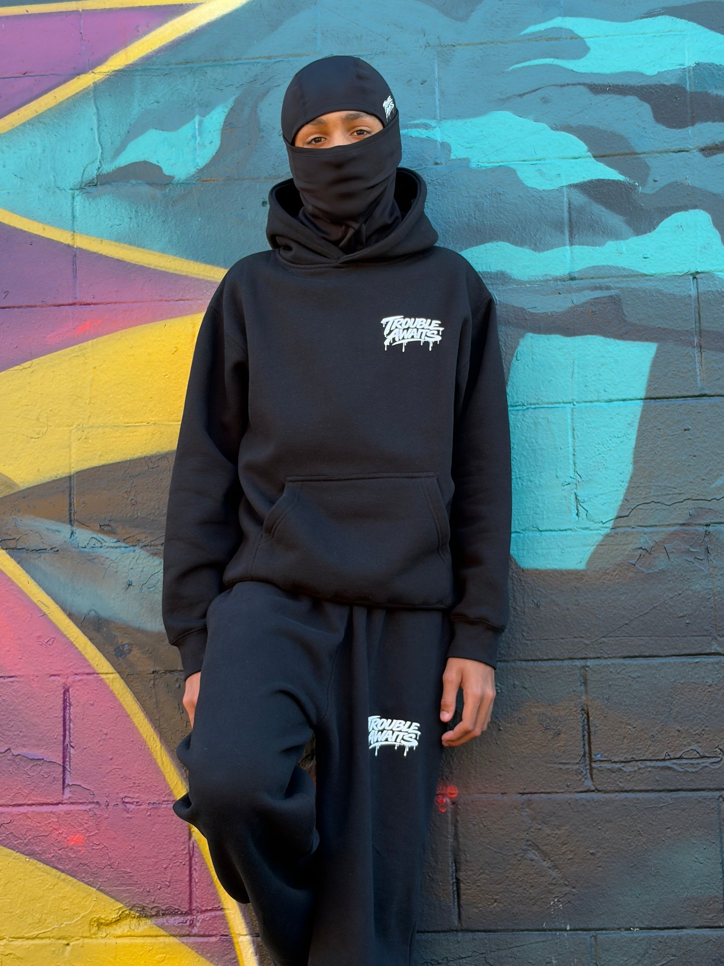 This image shows a person posing against a vibrant graffiti mural painted on a textured brick wall. The mural features large abstract shapes and arrows in bright yellow, purple, teal, and black, creating a bold and colorful urban backdrop. The person is positioned slightly to the side, leaning against the wall with one leg bent and foot resting against it. They are wearing a coordinated black streetwear outfit consisting of a pullover hoodie and matching sweatpants