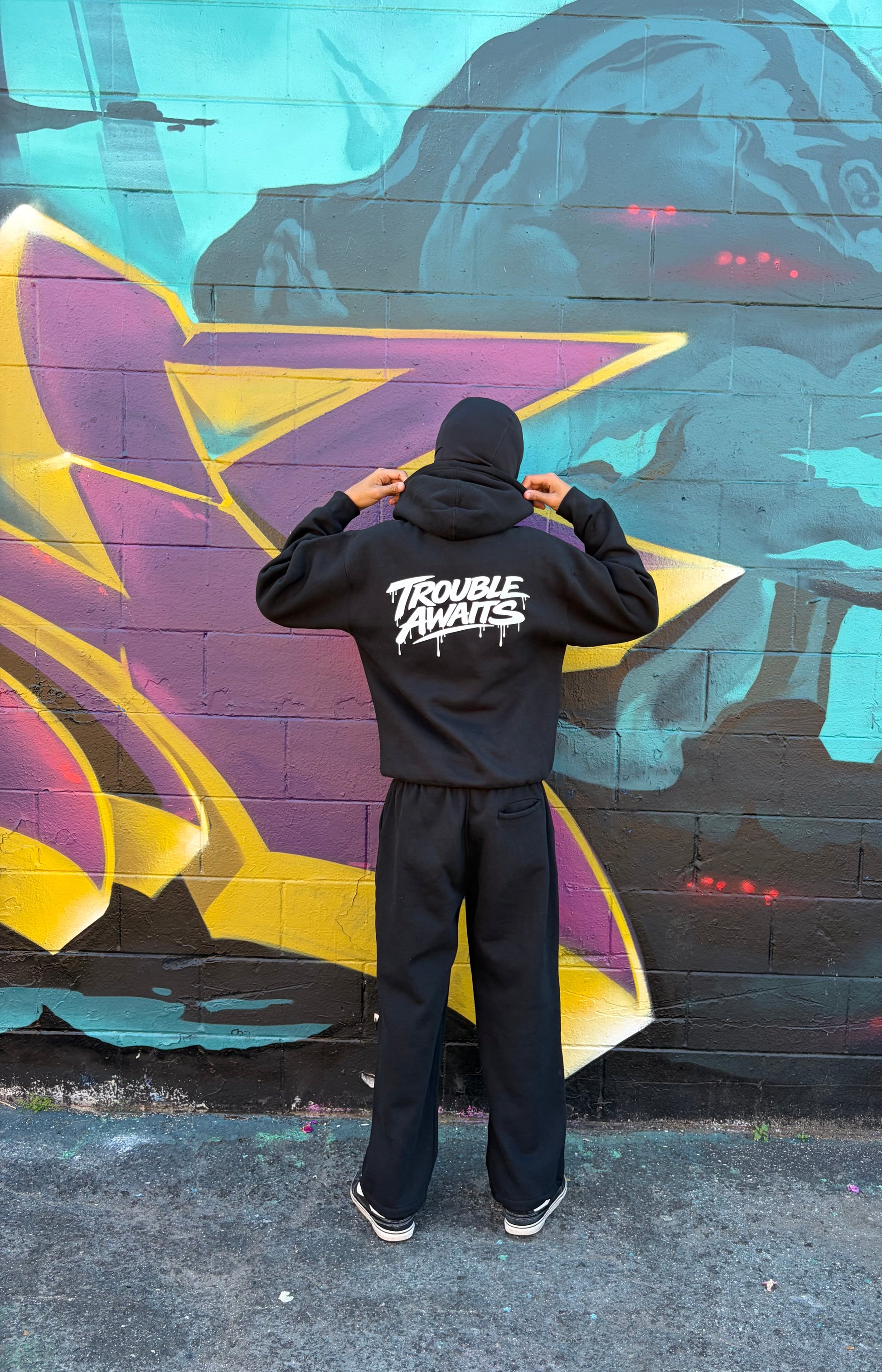 Person standing with their back to the camera wearing a black hoodie and matching black sweatpants set. The hoodie features a large white “Trouble Awaits” graphic printed across the back in a dripping, graffiti-style font. The person is holding the sides of the hood up with both hands. The sweatpants have a relaxed, straight-leg fit with a slightly baggy silhouette. They are standing in front of a colorful graffiti mural with bold shapes in teal, purple, yellow, and black painted on a brick wall. 