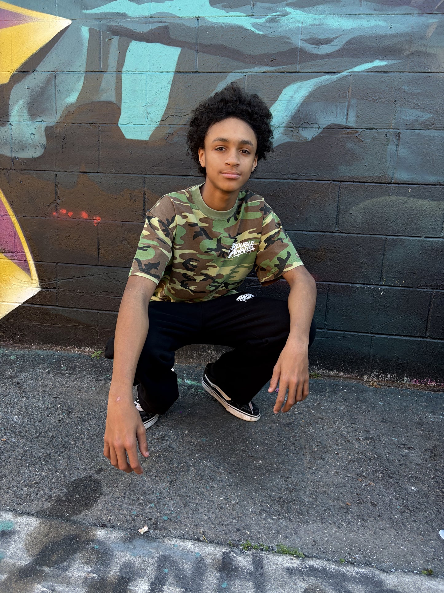 A young person is crouching outdoors on a concrete surface in front of a painted brick wall covered in graffiti art. They are wearing a short-sleeve T-shirt with a woodland camouflage pattern in green, brown, tan, and black. On the upper left chest of the shirt is a small white “Trouble Awaits” logo in a graffiti-style font with a subtle dripping effect. They are also wearing loose black pants and black low-top sneakers with white soles. The person is posed with one knee bent and one arm resting casually 