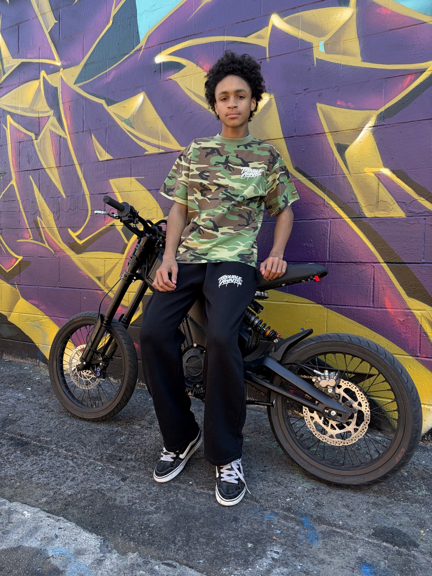 A young person is seated on a black electric bike parked on a concrete surface in front of a colorful graffiti-covered brick wall. The graffiti features bold shapes and angular lettering in purple, yellow, and blue tones. The person is wearing a short-sleeve T-shirt with a woodland camouflage pattern in green, brown, tan, and black. On the upper left chest is a small white “Trouble Awaits” logo in a graffiti-style font with a subtle dripping effect. They are also wearing loose black pants.
