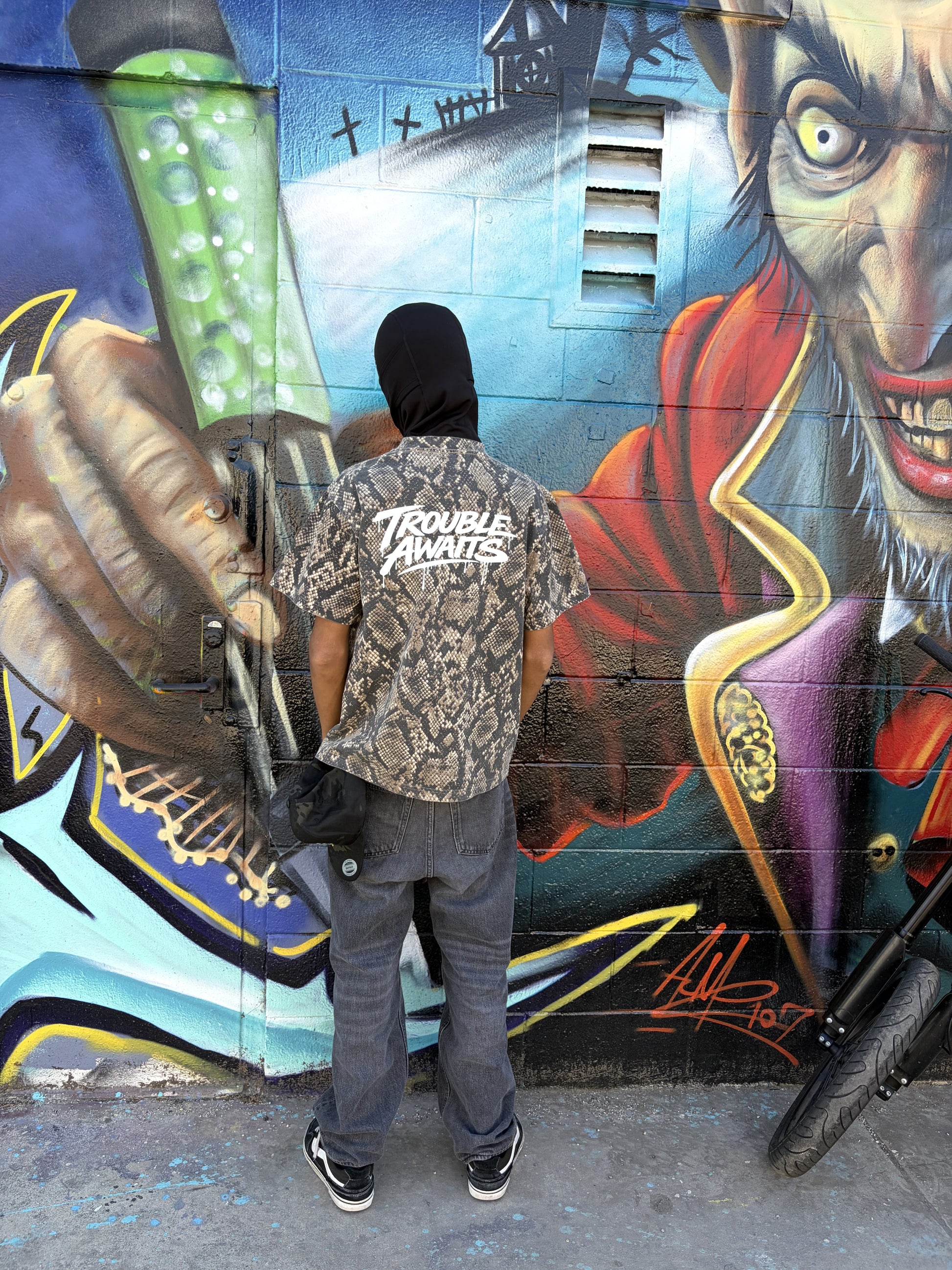 A person stands outdoors in front of a brightly colored graffiti mural painted on a textured brick wall. The artwork features bold shapes and layered colors, including light blue, red, yellow, and black outlines, with stylized lettering and abstract forms. The ground is rough concrete with small debris and a white bag visible near the lower edge.

The person is facing the camera in a relaxed pose, leaning slightly back against the wall with one leg bent and the foot resting on the wall behind them. 
