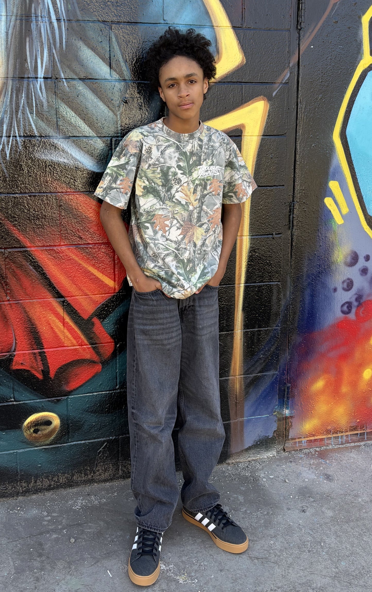 A person stands outdoors in front of a textured brick wall covered in colorful graffiti artwork. The mural includes bold shapes and blended colors such as black, red, yellow, and blue, with layered spray-painted elements and abstract forms. The ground is rough concrete with small debris scattered near the base of the wall.

The person is facing the camera in a relaxed stance, leaning slightly back against the wall with both hands in their pockets. They are wearing an oversized, boxy short-sleeve T-shirt 