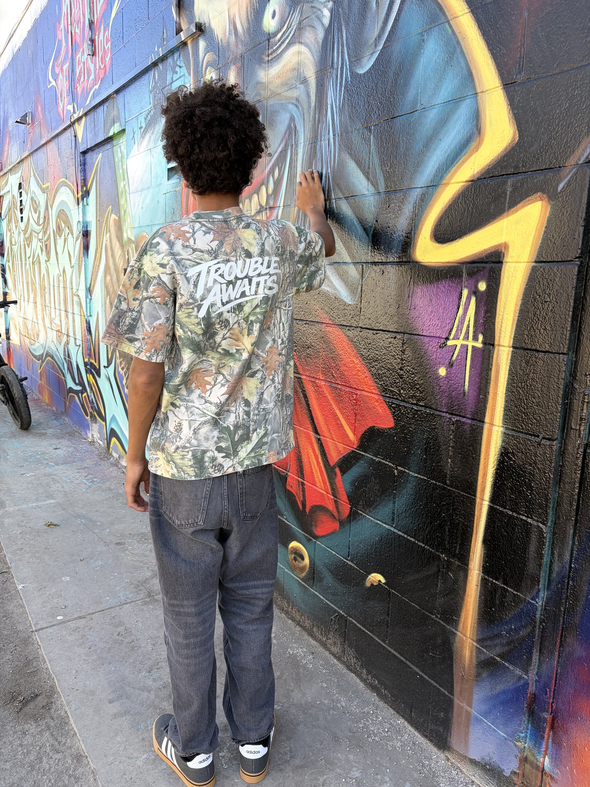 A person is standing outdoors beside a brick wall covered in vibrant graffiti artwork. The mural features bold, layered colors including black, blue, red, yellow, and purple, with stylized shapes and abstract figures. The ground is a concrete sidewalk with small marks and debris typical of an urban setting.

The person is facing away from the camera, with one hand lightly touching the wall. They are wearing an oversized, boxy short-sleeve T-shirt with a realistic woodland camouflage pattern 