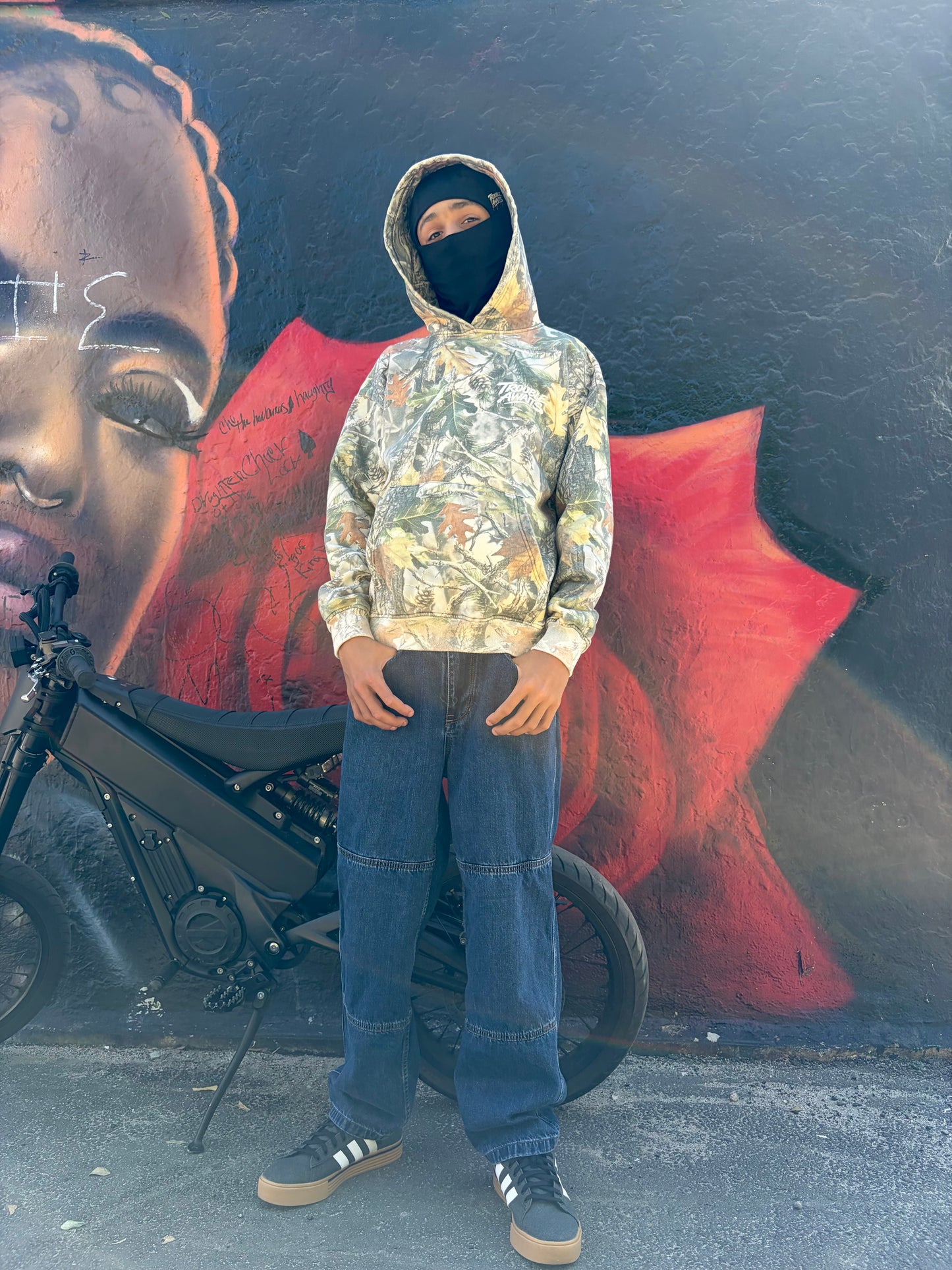 This image shows a person standing outdoors against a large painted mural on a textured wall. The mural features a stylized portrait of a face on the left side and bold red shapes against a dark background. The person is centered in the frame, facing forward with a relaxed posture and arms resting at their sides. They are wearing a camouflage-pattern hoodie with a forest-inspired design of leaves and branches in green, brown, and gray tones. A small white “Trouble Awaits” logo in a graffiti-style font