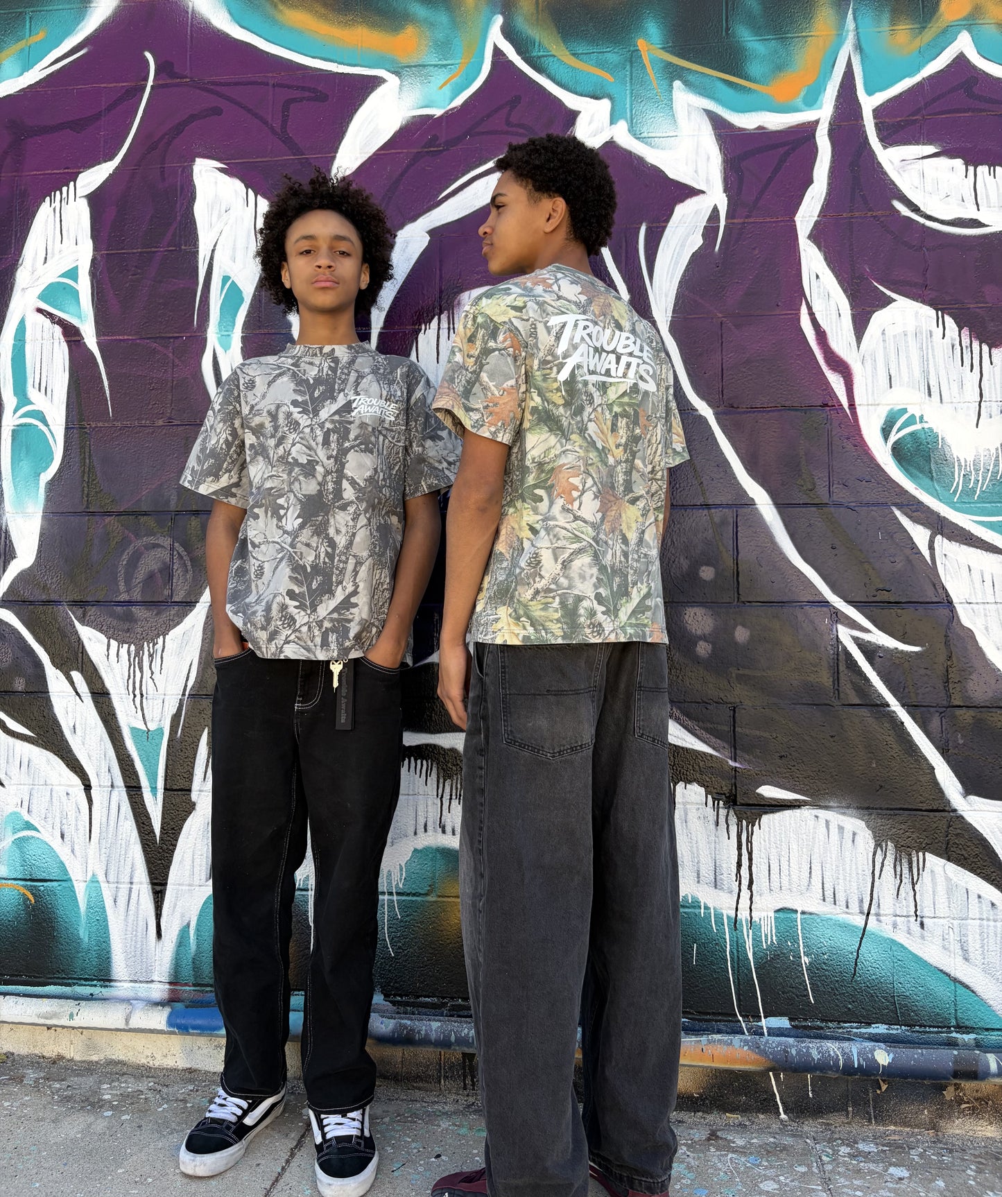 Two people stand in front of a graffiti-covered brick wall featuring bold, layered artwork in purple, teal, white, and black, with dripping paint effects and sharp, abstract shapes. The ground is a worn concrete surface, adding to the urban street setting.

The person on the left faces the camera directly with a neutral expression and relaxed posture, hands in their pockets. They are wearing an oversized, boxy short-sleeve T-shirt with a gray-toned woodland camouflage pattern 