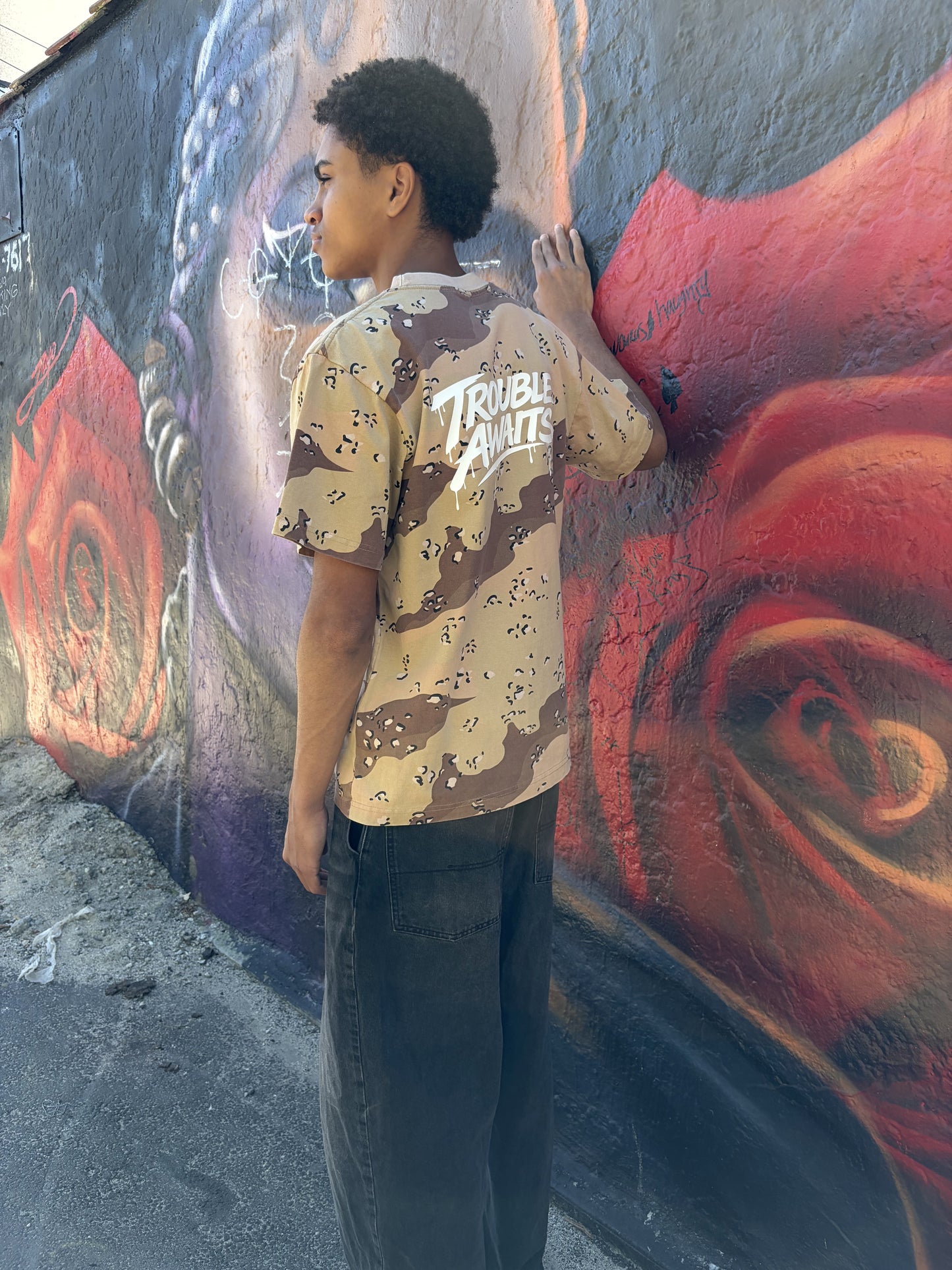 A person stands outdoors beside a wall covered in large, colorful graffiti artwork. The mural features a mix of dark gray, black, red, and purple tones with layered shapes, abstract forms, and spray-painted tags. The ground is rough concrete with small debris scattered near the base of the wall.

The person is shown from a three-quarter back view, facing the wall with one hand lightly touching it while their head is turned slightly to the side. They are wearing an oversized, boxy short-sleeve T-shirt 