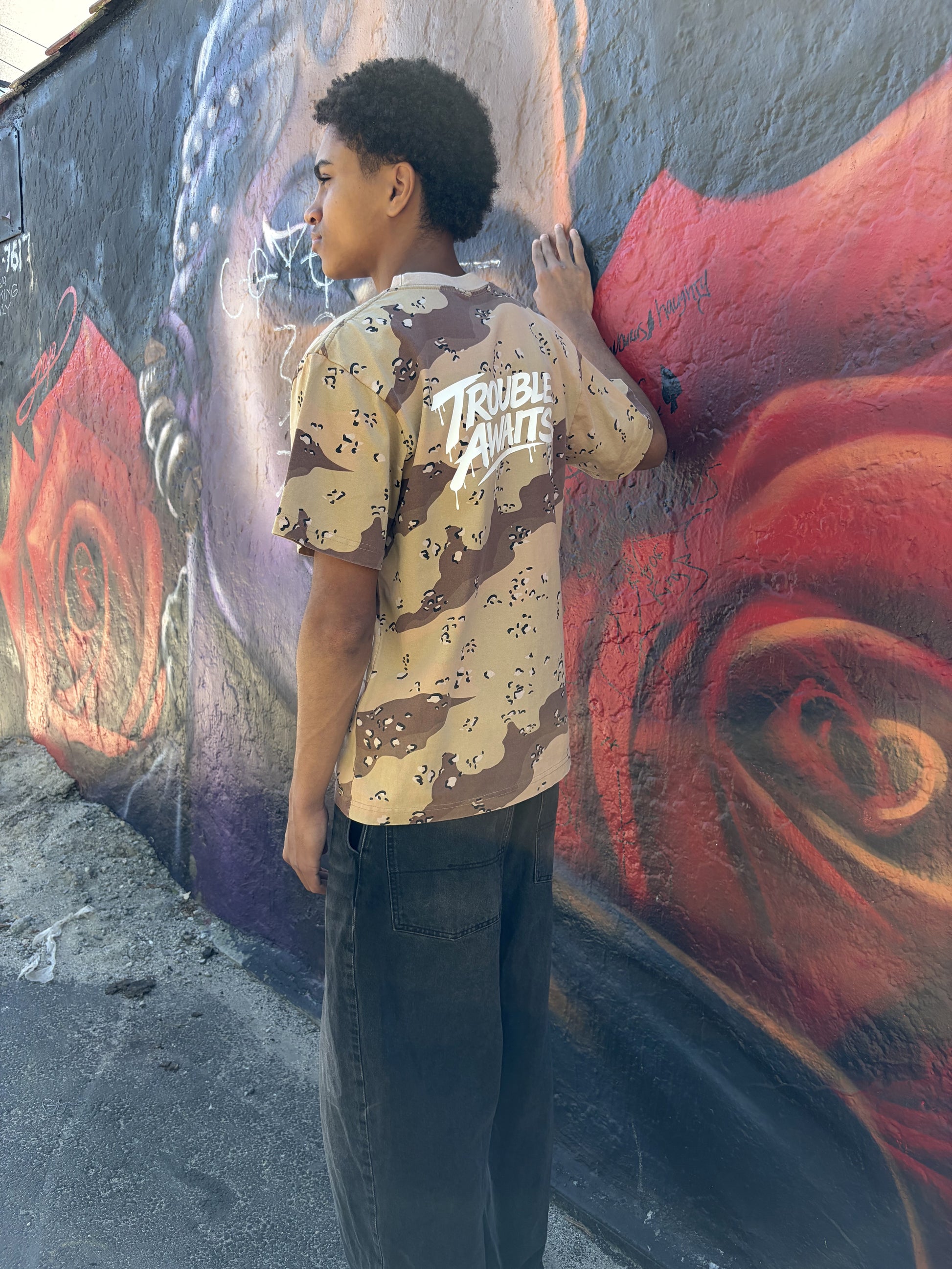 A person stands outdoors beside a wall covered in large, colorful graffiti artwork. The mural features a mix of dark gray, black, red, and purple tones with layered shapes, abstract forms, and spray-painted tags. The ground is rough concrete with small debris scattered near the base of the wall.

The person is shown from a three-quarter back view, facing the wall with one hand lightly touching it while their head is turned slightly to the side. They are wearing an oversized, boxy short-sleeve T-shirt 