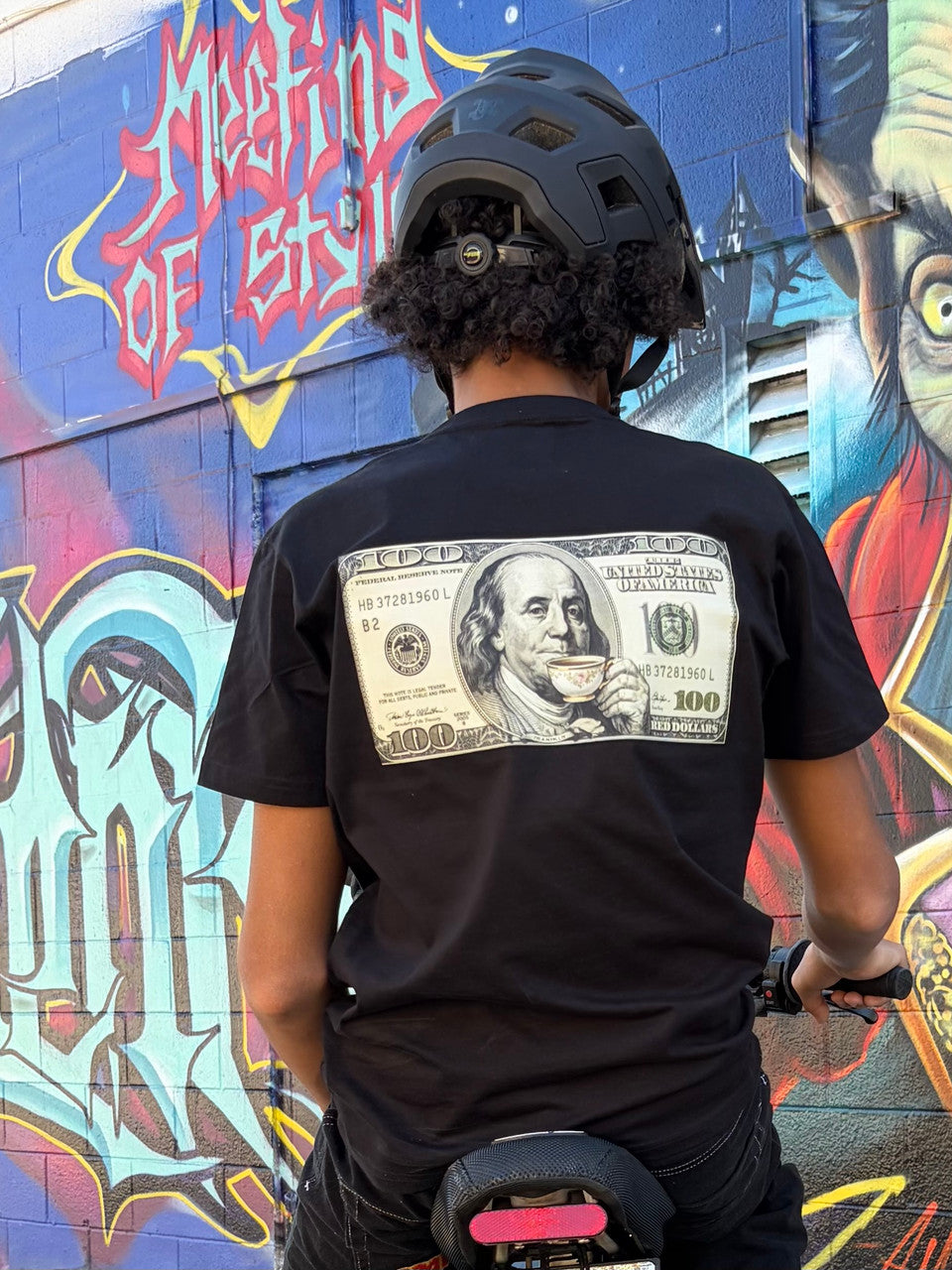 A person viewed from behind wearing a black t-shirt with a large graphic on the upper back showing a stylized $100 bill with Benjamin Franklin sipping tea from a small teacup. The person is wearing a black helmet and appears to be sitting on a Talaria XXX electric motorcyle. The background features a brightly colored graffiti mural painted on a wall with bold lettering and vibrant colors.