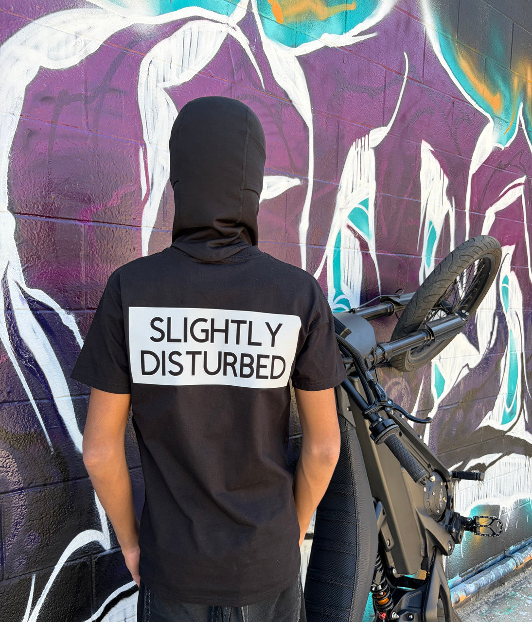 A person viewed from behind wearing a black t-shirt with a large white rectangular graphic on the upper back that reads “SLIGHTLY DISTURBED” in bold black capital letters. The person is also wearing a black head covering and stands next to a electric Talaria XXX bike. The background features a colorful graffiti mural with abstract shapes and bold colors painted on a wall.