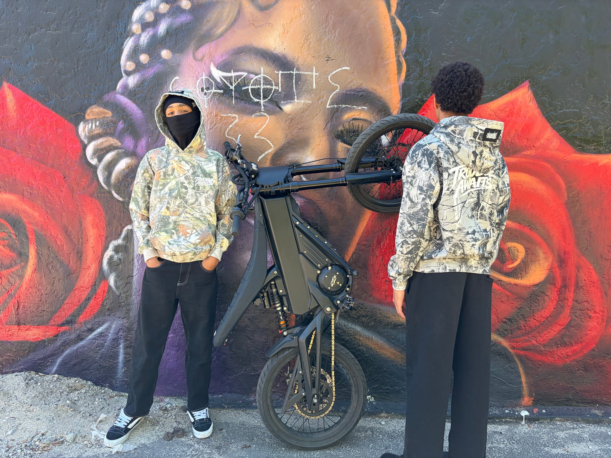This image shows two people posing outdoors in front of a large mural painted on a textured wall. The mural features a stylized face with braided hair and bold red shapes against a dark background. A black electric-style bike is positioned between them, with its frame and front wheel visible.

The person on the left faces the camera and wears a forest camouflage hoodie with a pattern of leaves and branches in green, brown, and tan tones. The hoodie has a small white “Trouble Awaits” logo on the left chest. 