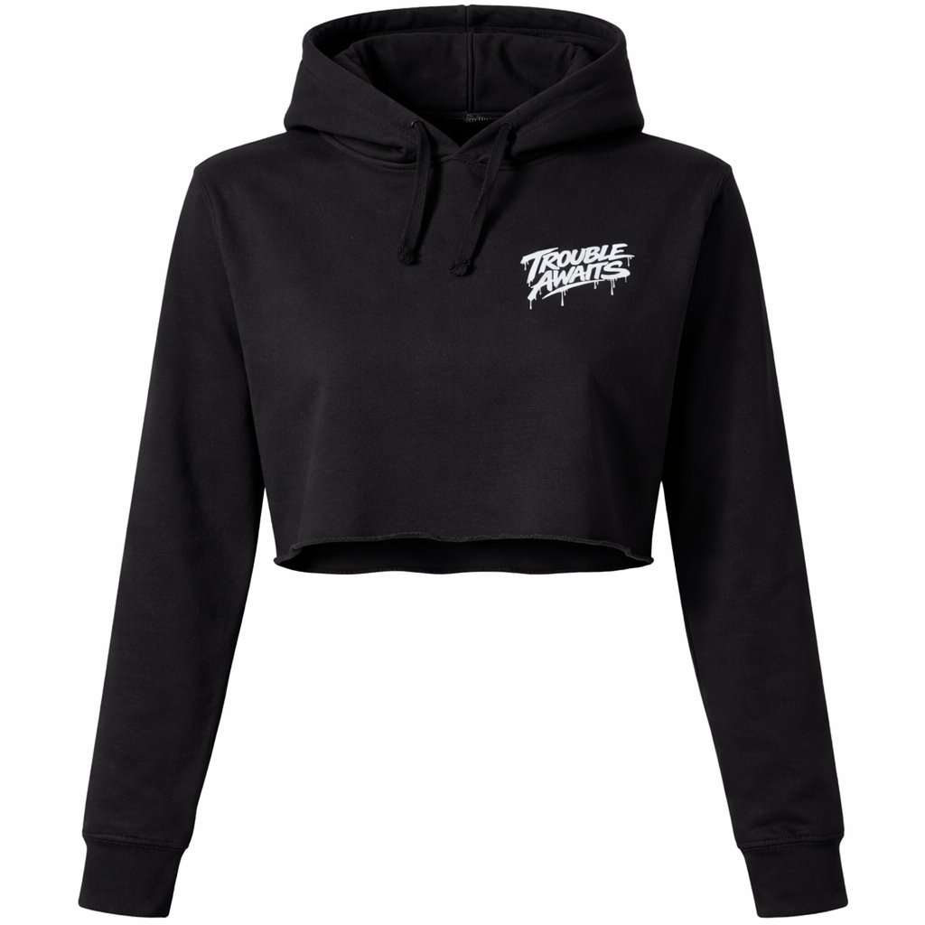 A product image shows the front of a black cropped hoodie displayed against a plain white background. The hoodie features a relaxed fit with long sleeves, a hood, and black drawstrings at the neckline. The bottom hem is cropped with a raw, unfinished edge, giving it a slightly distressed look. On the upper left chest (viewer’s right side), there is a small white “Trouble Awaits” logo in a bold graffiti-style font with subtle dripping paint details. The rest of the hoodie is plain black. 