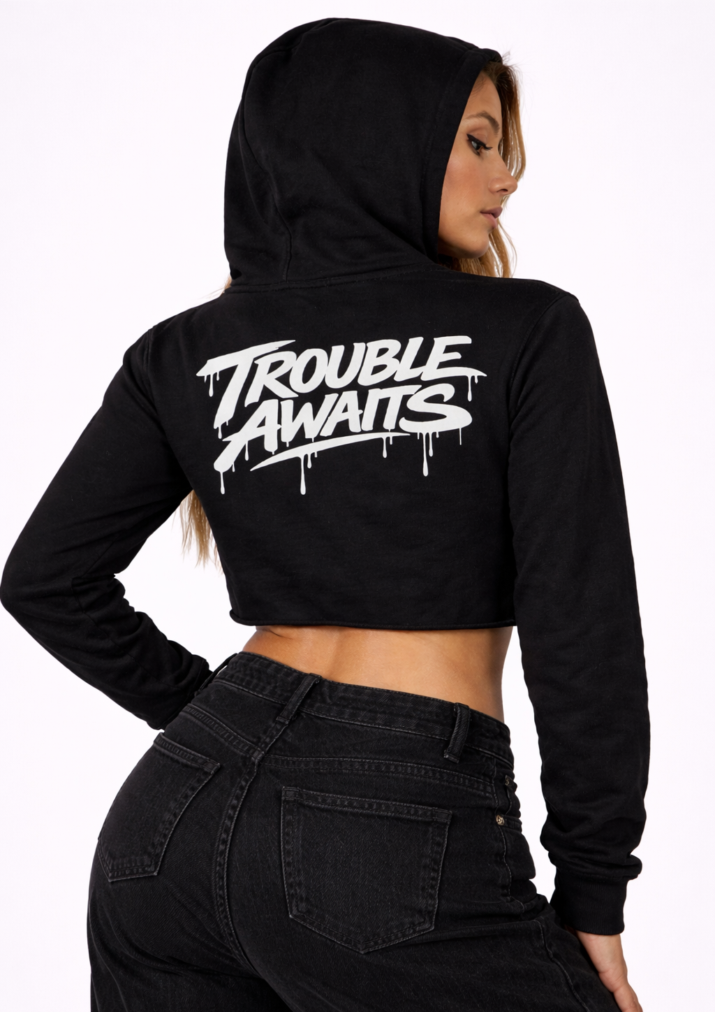 A studio photo shows a woman from behind, slightly turned to the side, modeling a black cropped hoodie. The hoodie has a relaxed fit with long sleeves and a hood up, and features a raw, unfinished bottom hem with no ribbed band. Centered across the upper back is a large white “Trouble Awaits” logo in a bold graffiti-style font with dripping paint details. The cropped cut exposes a small portion of her lower back above high-waisted black jeans. The background is plain white with even lighting