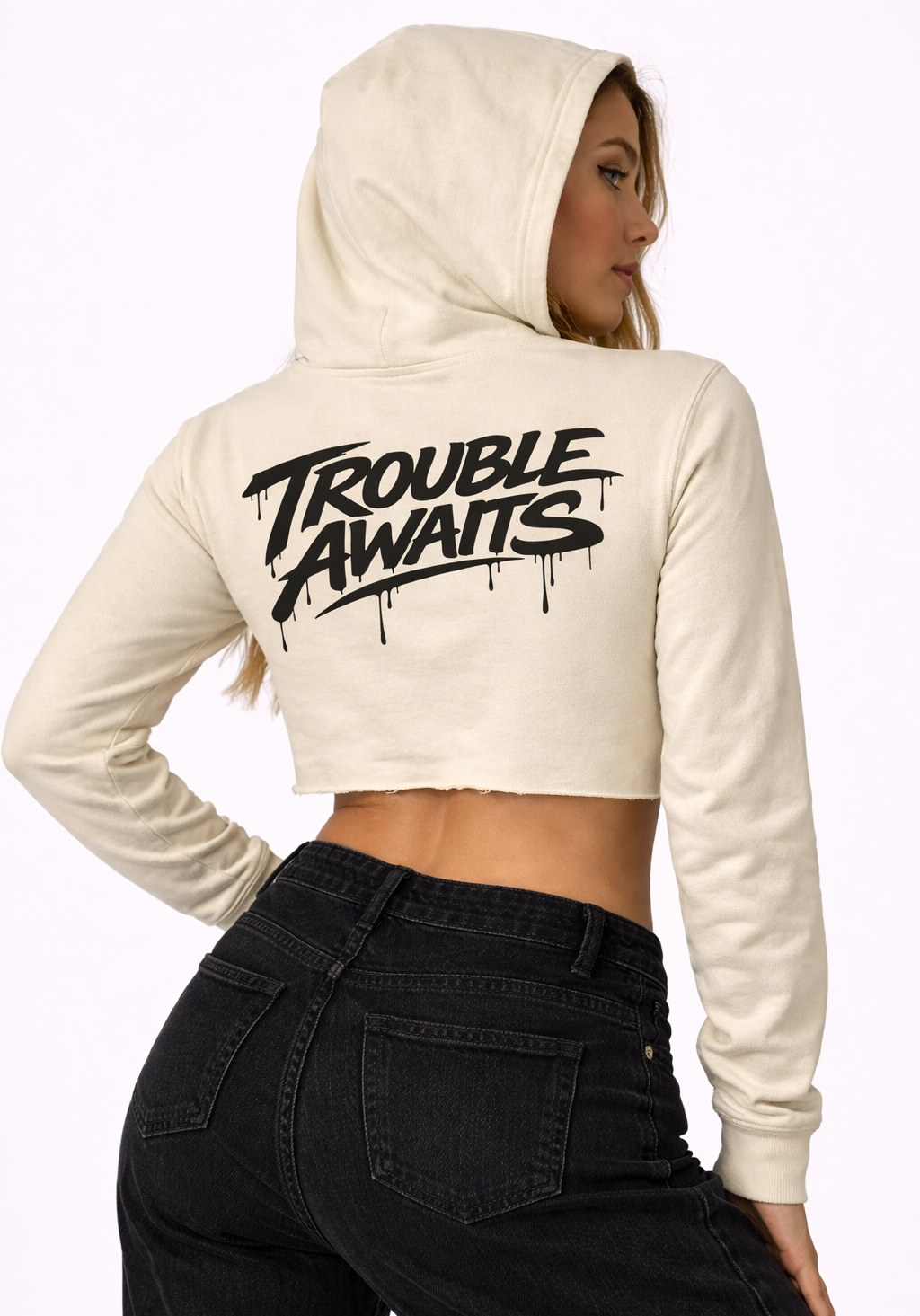 A woman is shown from a three-quarter back angle wearing a cropped cream/beige hoodie. The hoodie has a fitted yet slightly relaxed shape with long sleeves and a raw-cut hem that sits above the waist. The hood is up, framing her head, and her hair is partially visible along the side of her face. Across the upper back is a large black “Trouble Awaits” graphic in a bold graffiti-style script with dripping paint details. She pairs the hoodie with high-waisted black jeans. The background is plain