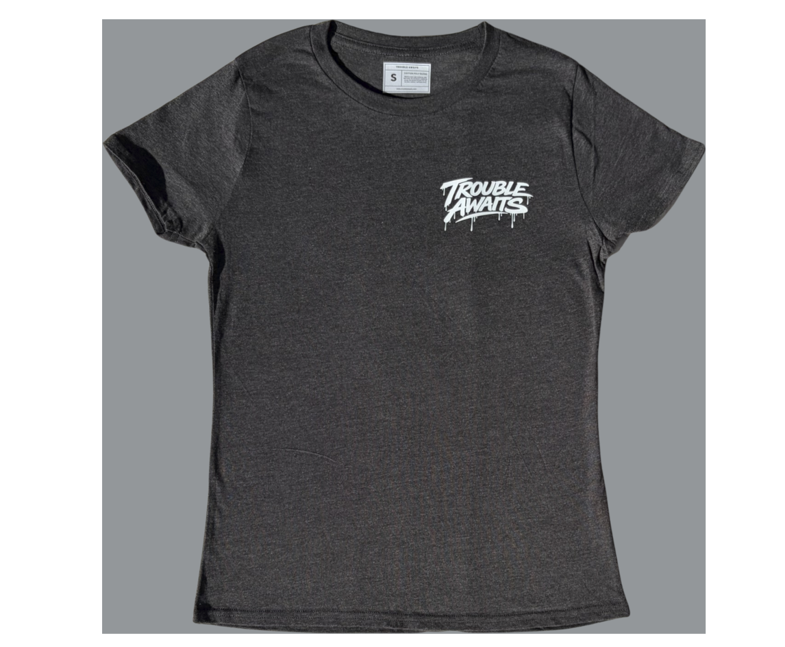 A front view of a women’s fitted T-shirt in heather gray, displayed against a neutral gray background. The shirt has a slightly contoured, tapered silhouette with short sleeves and a crew neckline. A small printed size label is visible inside the collar.

On the left chest area, there is a small white “Trouble Awaits” graphic in a graffiti-style font with subtle dripping paint details, creating a bold streetwear look. The design sits high on the chest and contrasts clearly against the darker heather gray 