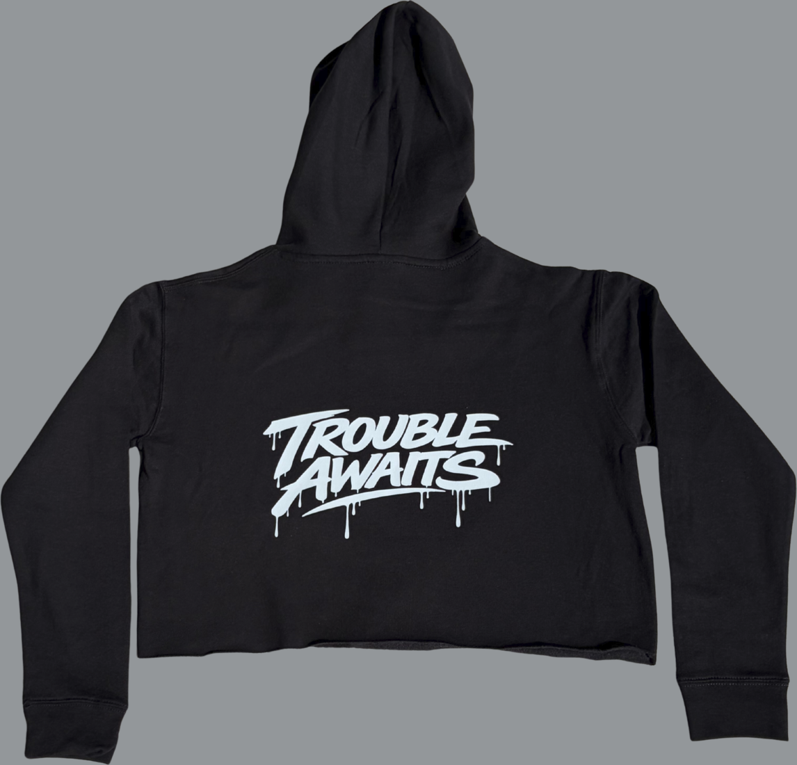 A product image shows the back of a black cropped hoodie laid flat against a neutral gray background. The hoodie has long sleeves and a hood, with a short, cropped body and a raw, unfinished bottom hem (no ribbed band). Centered across the upper back is a large white “Trouble Awaits” logo in a bold graffiti-style font with dripping paint details. The fabric appears smooth and slightly structured, with subtle folds visible along the sleeves and body. 