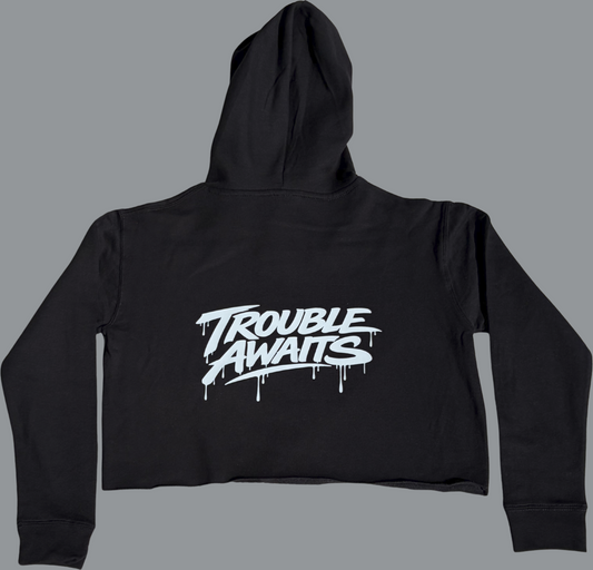 A product image shows the back of a black cropped hoodie laid flat against a neutral gray background. The hoodie has long sleeves and a hood, with a short, cropped body and a raw, unfinished bottom hem (no ribbed band). Centered across the upper back is a large white “Trouble Awaits” logo in a bold graffiti-style font with dripping paint details. The fabric appears smooth and slightly structured, with subtle folds visible along the sleeves and body. 