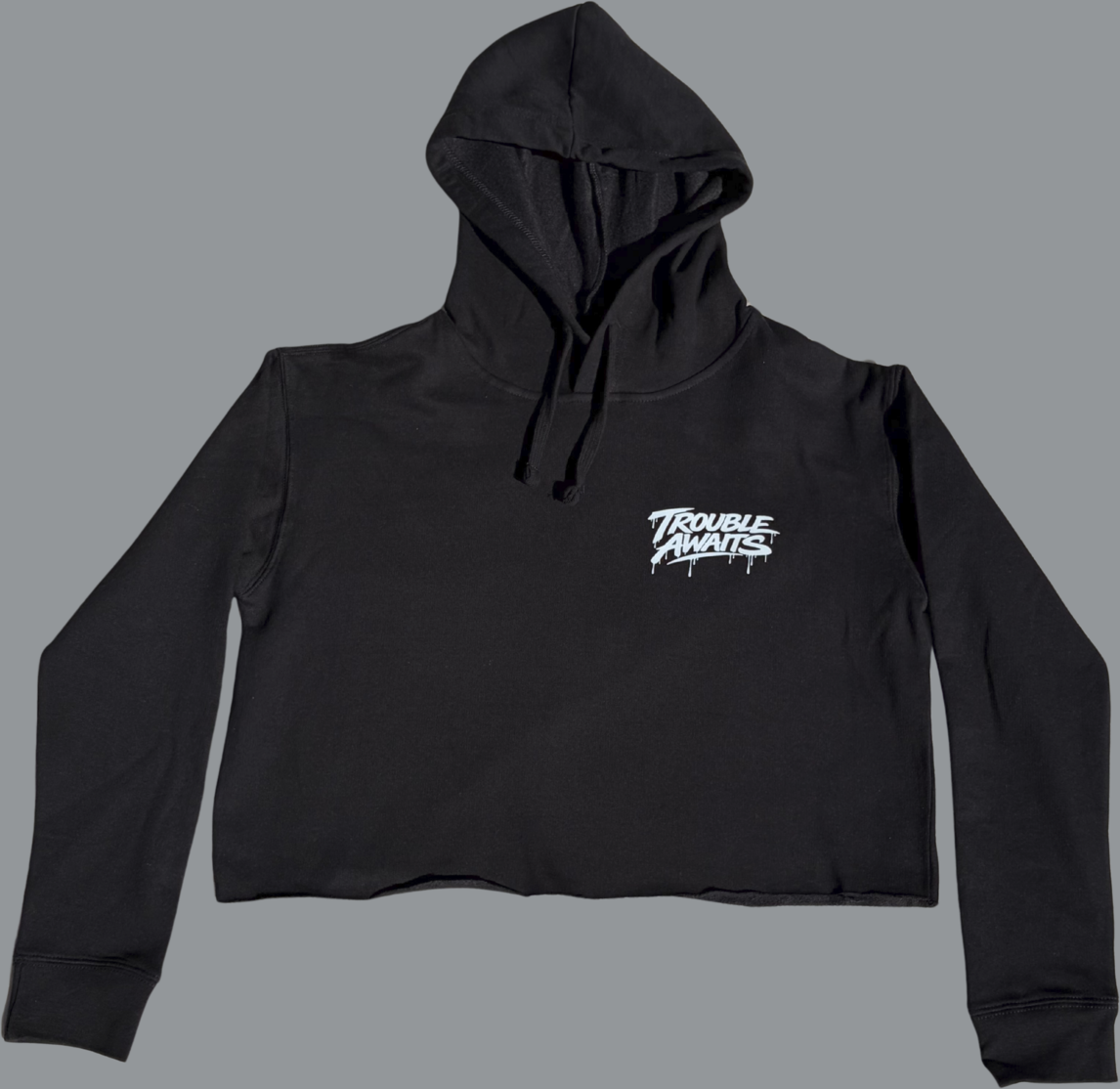 A product image shows the front of a black cropped hoodie laid flat against a neutral gray background. The hoodie features a hood with drawstrings and long sleeves. The body is cropped with a raw, unfinished bottom hem, giving it a slightly rugged edge. On the upper left chest (viewer’s right side), there is a small white “Trouble Awaits” logo in a graffiti-style font with subtle dripping paint details. The rest of the hoodie is plain black. The fabric appears soft and slightly structured