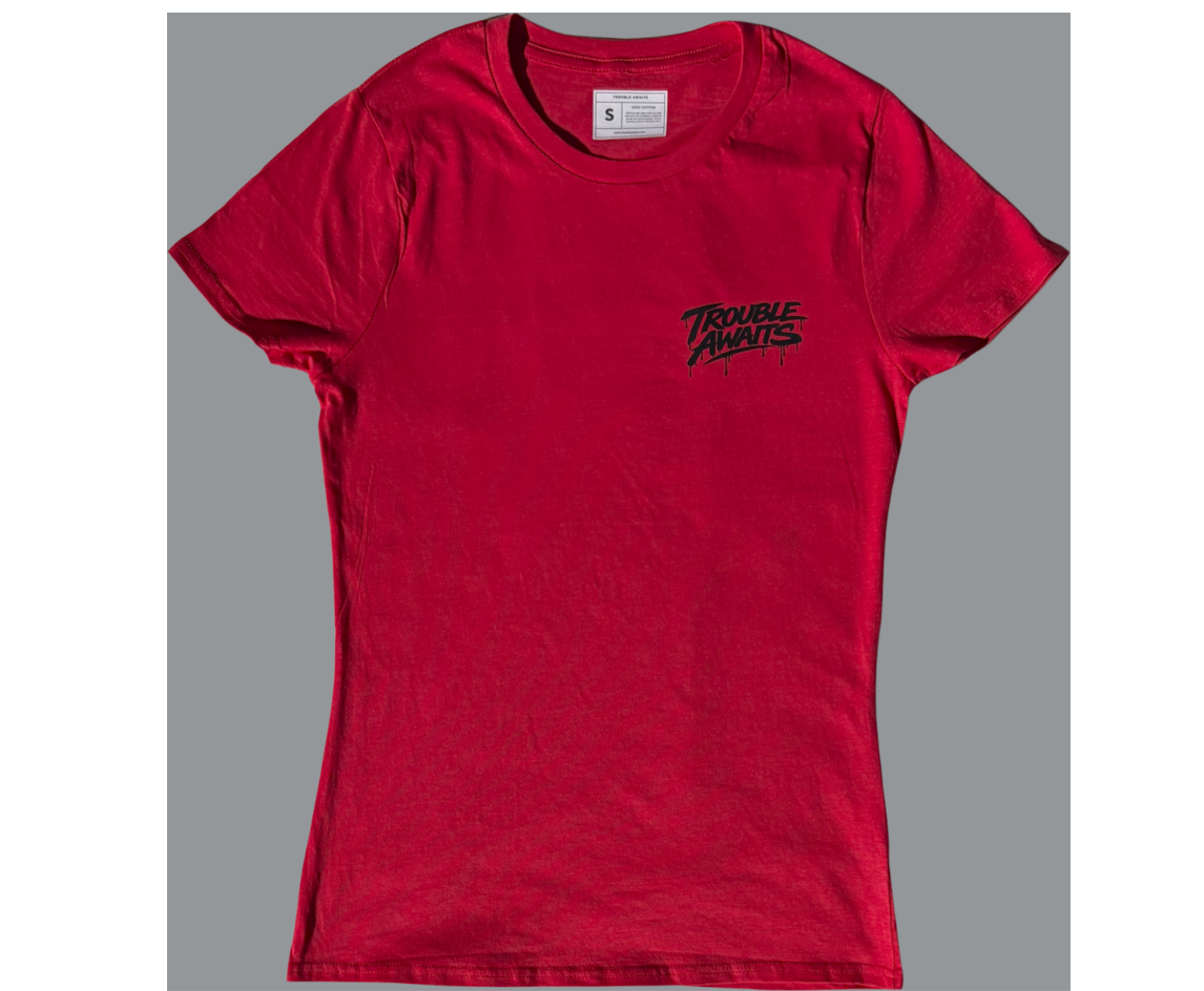 A front view of a women’s lightweight T-shirt in a rich red color, displayed against a neutral gray background. The shirt features a slightly fitted, contoured silhouette with short sleeves and a crew neckline. A small printed label is visible inside the collar.

On the left chest area, there is a small black “Trouble Awaits” graphic in a graffiti-style font with subtle dripping paint details, giving it a streetwear-inspired look. The print is positioned higher on the chest and contrasts clearly 