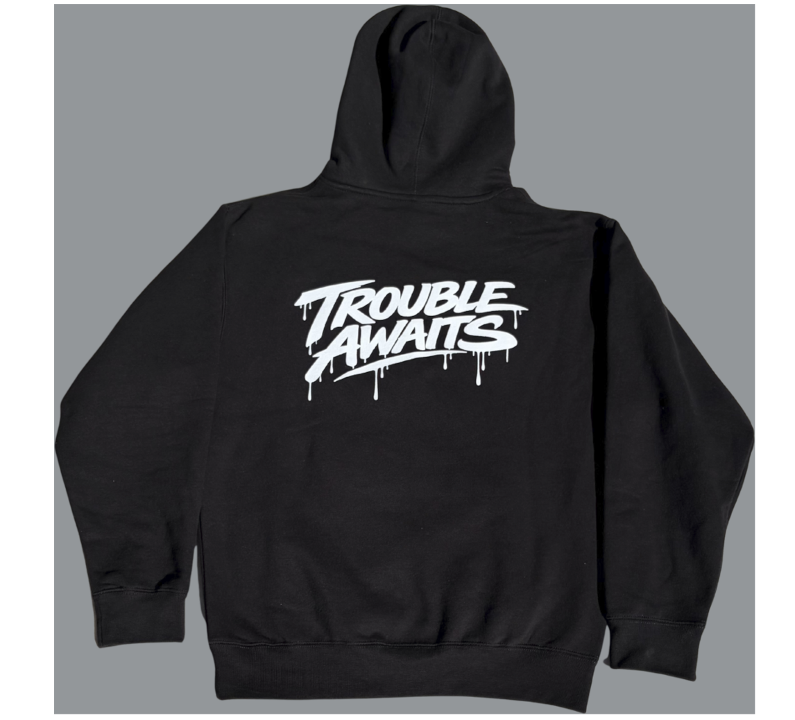 this image shows the back view of a black pullover hoodie laid flat against a plain gray background. The hoodie features a large white graphic centered across the upper back that reads “Trouble Awaits” in a stylized graffiti-inspired font. The lettering has a hand-painted look with drip effects extending downward, giving it a street-art aesthetic. The hood is visible at the top, and the garment appears to have a standard relaxed fit with long sleeves, ribbed cuffs, and a ribbed waistband.
