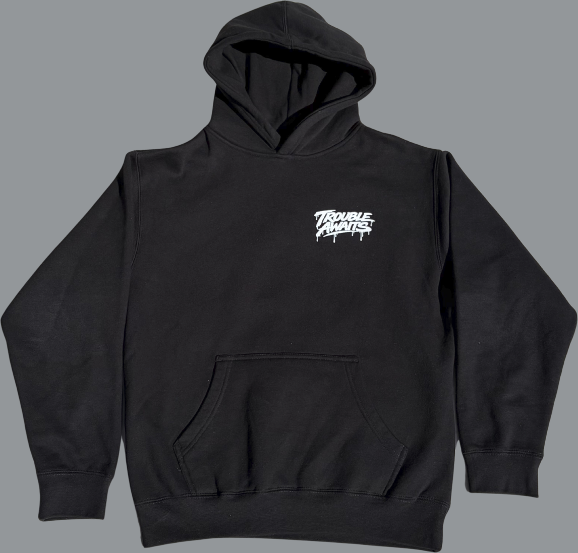 This image shows the front view of a black pullover hoodie laid flat against a plain gray background. The hoodie has a clean, minimal design with a small white graphic on the left chest that reads “Trouble Awaits” in a stylized graffiti-inspired font, featuring slight paint-drip accents beneath the lettering. The garment includes a spacious front kangaroo pocket, a hood with visible drawstrings, long sleeves, and ribbed cuffs and waistband. The overall fit appears relaxed and casual