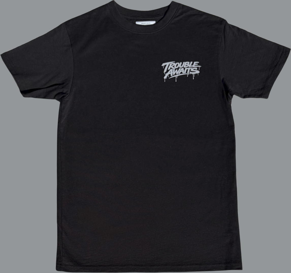 Black short sleeve t-shirt featuring a small light grey graphic printed on the left chest. The design reads “Trouble Awaits” in stylized lettering with a dripping paint effect beneath the letters. The rest of the shirt is plain black with no additional visible graphics.