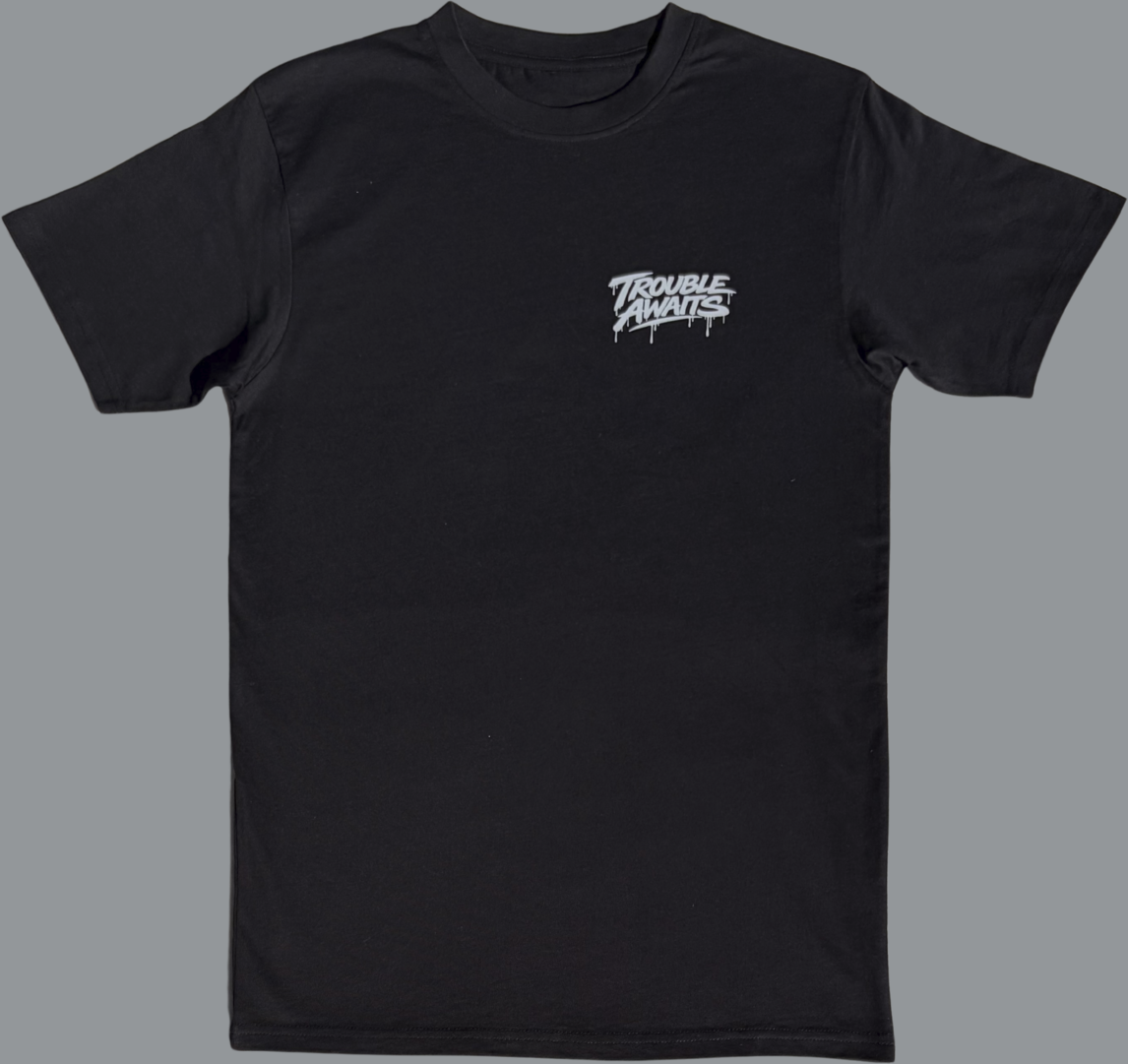 Black short sleeve t-shirt featuring a small light graphic printed on the left chest. The design reads “Trouble Awaits” in stylized lettering with a dripping paint effect beneath the letters. The rest of the shirt is plain black with no additional visible graphics.