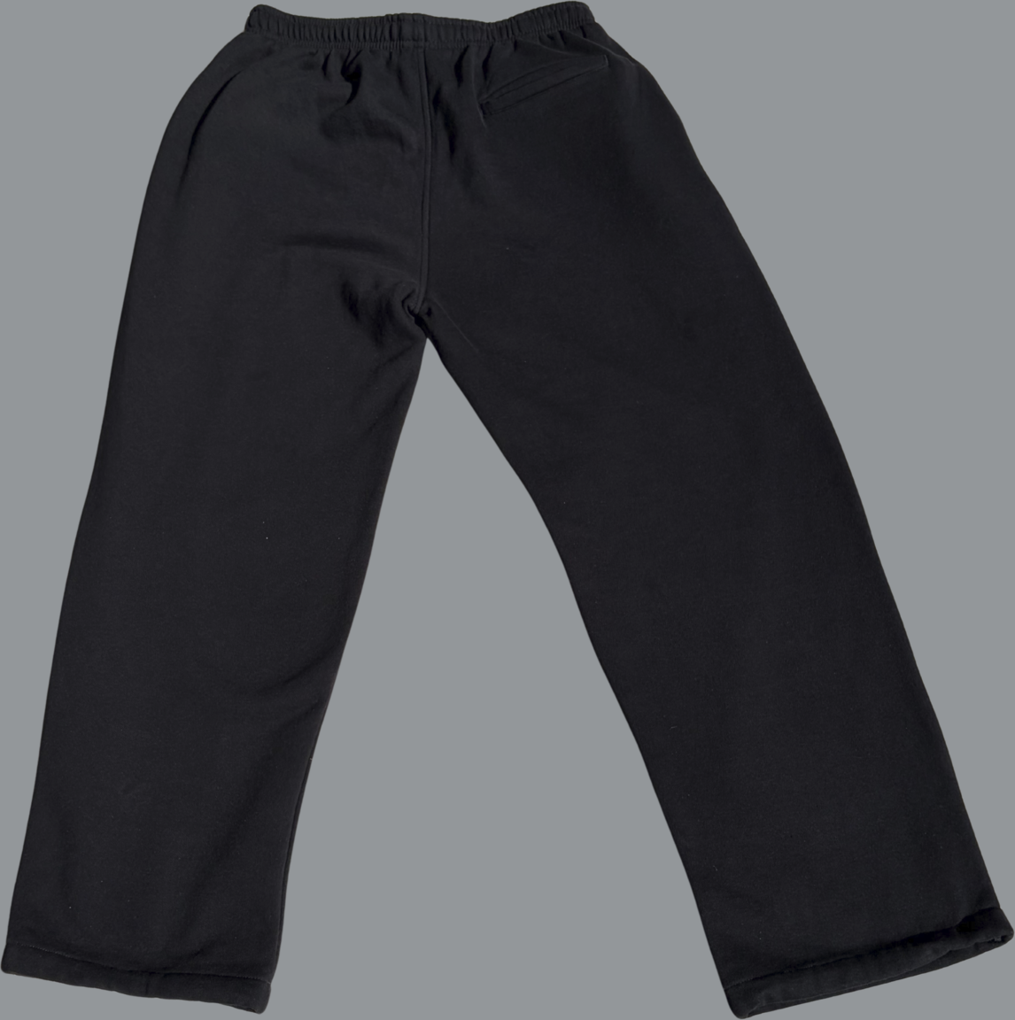 Back view of black baggy sweatpants laid flat on a light gray background. The pants feature a relaxed, straight-leg fit with wide openings and no cuffs, and an elastic waistband at the top. A single back pocket is visible on the right side. The fabric appears soft and slightly heavy, with subtle creases that highlight the loose, oversized silhouette.