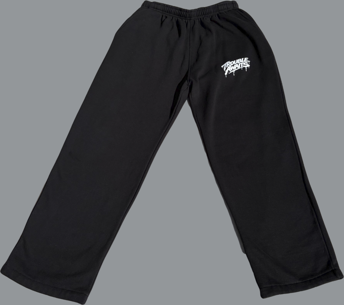 Front view of black baggy sweatpants laid flat on a light gray background. The pants feature a relaxed, straight-leg fit with wide openings and no cuffs, and an elastic waistband at the top. A small white “Trouble Awaits” logo is printed on the upper right thigh. The fabric appears soft and slightly heavy, with natural folds that emphasize the loose, oversized silhouette.