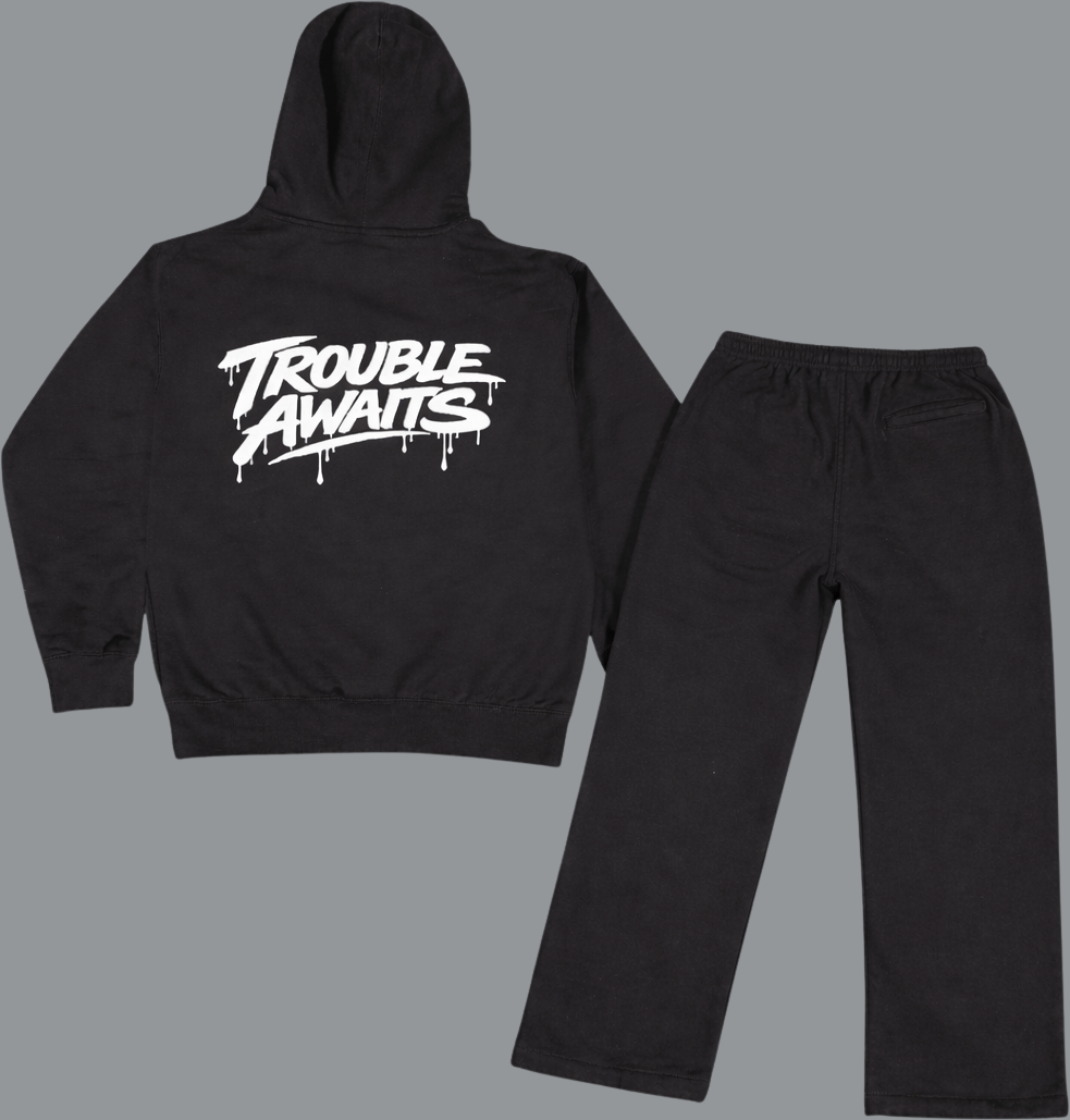 Flat lay of a black hoodie and matching black sweatpants shown from the back on a light gray background. The hoodie features a large white “Trouble Awaits” graphic across the back in a dripping, graffiti-style font, with a hood and ribbed hem and cuffs. The sweatpants have a relaxed, straight-leg fit with an elastic waistband and a single back pocket visible on the right side. The legs appear slightly cropped in the layout, emphasizing a baggy, wide-leg silhouette rather than a long, tapered fit.