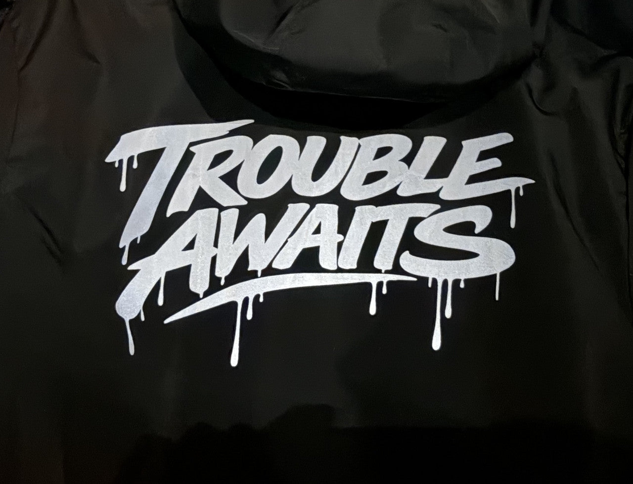 A close-up view of the back of a black windbreaker jacket focuses on a bold silver graphic reading “Trouble Awaits.” The lettering is in a graffiti-inspired script with thick strokes and stylized edges. Paint-drip details extend downward from several letters, giving it a street art aesthetic. The print sits centered on the upper back beneath the hood. The fabric appears smooth and slightly reflective with visible creases, suggesting a lightweight, water-resistant material.