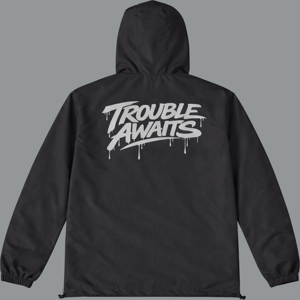 A lightweight black hooded windbreaker jacket is shown from the back against a neutral gray background. The jacket has a relaxed, slightly oversized fit with a loose body, elasticated cuffs, and a cinched hem. The hood is up and structured, with subtle seam lines visible. The fabric appears smooth with light wrinkling, suggesting a thin, water-resistant material. Centered across the upper back is a bold silver graphic reading “Trouble Awaits” in a stylized graffiti-inspired font