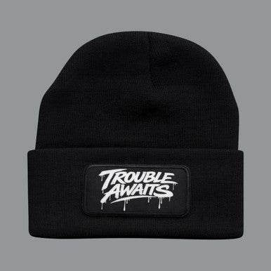 A black knit beanie is shown from the front against a neutral background. The beanie has a ribbed texture and a folded cuff, giving it a snug, classic fit. Centered on the cuff is a rectangular patch with the “Trouble Awaits” logo in bold white graffiti-style lettering, featuring paint-drip details beneath the text. The fabric appears soft and slightly stretchy, with subtle variations in lighting that highlight the knit pattern. The overall design is minimal and streetwear-inspired