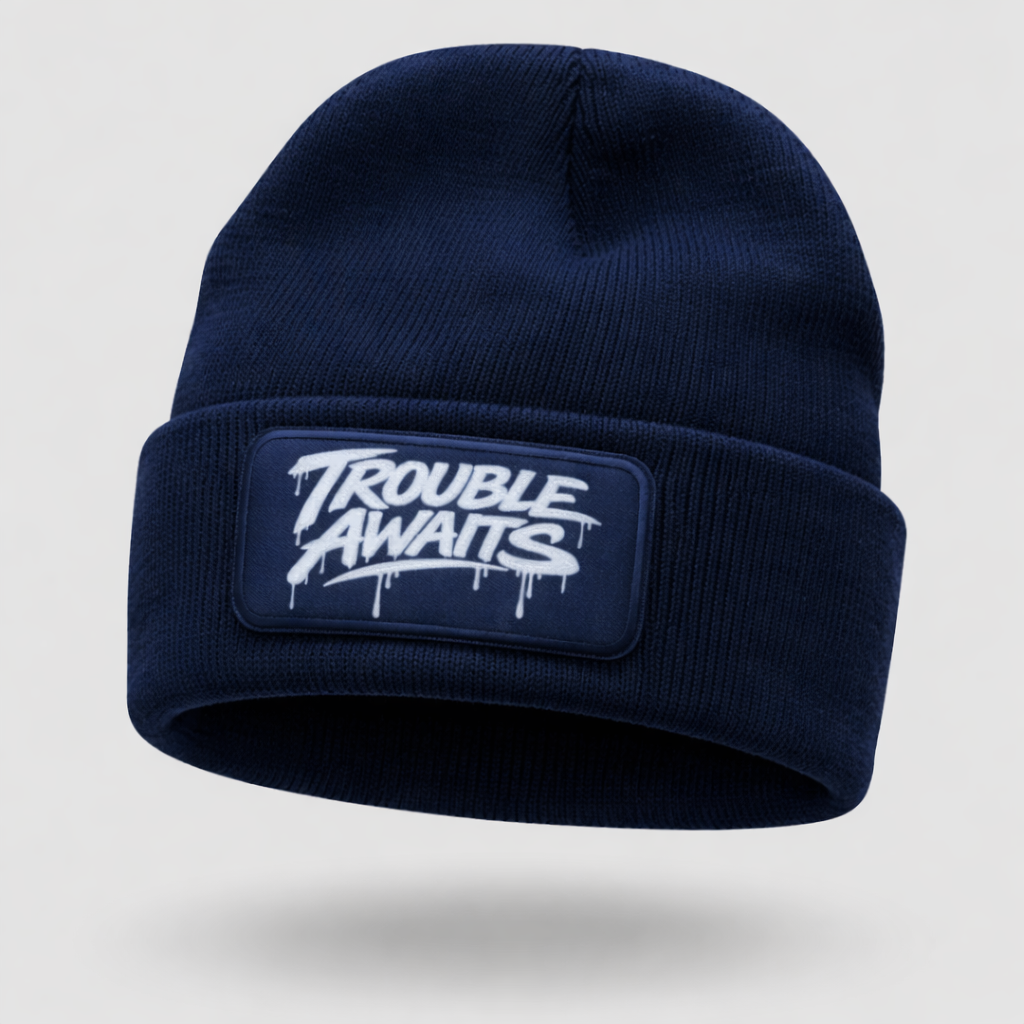 A black ribbed knit beanie is shown in a clean product-style image against a light gray background. The beanie has a folded cuff and a snug, rounded shape. Centered on the cuff is a rectangular black patch featuring the “Trouble Awaits” logo in bold white graffiti-style lettering with paint-drip details. The knit texture is clearly visible, giving the hat a soft, slightly stretchy appearance. The image has a subtle floating or ghost mannequin effect, with a faint shadow beneath the beanie 