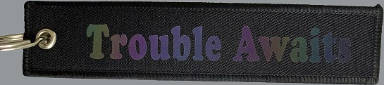 A rectangular black fabric key tag displayed against a neutral background. The tag has a textured, woven surface with stitched edges and a metal grommet on the left side, attached to a small metal keyring. Centered on the tag is the phrase “Trouble Awaits” in a clean, bold font. The lettering has a subtle holographic or color-shifting effect, reflecting tones of purple, green, and blue depending on the light. The overall design is minimal and durable, combining a soft fabric base with a reflective logo 