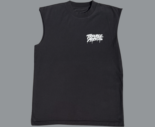 A sleeveless T-shirt (muscle tee) is displayed flat against a gray background, shown from the front. The shirt has a relaxed, boxy fit with wide arm openings and a crew neckline. The fabric appears smooth with subtle texture visible under even lighting.

The shirt is solid black, giving it a clean and minimal look. On the upper left chest, there is a small white “Trouble Awaits” logo in a graffiti-style font. The lettering is bold with stylized paint-drip details 